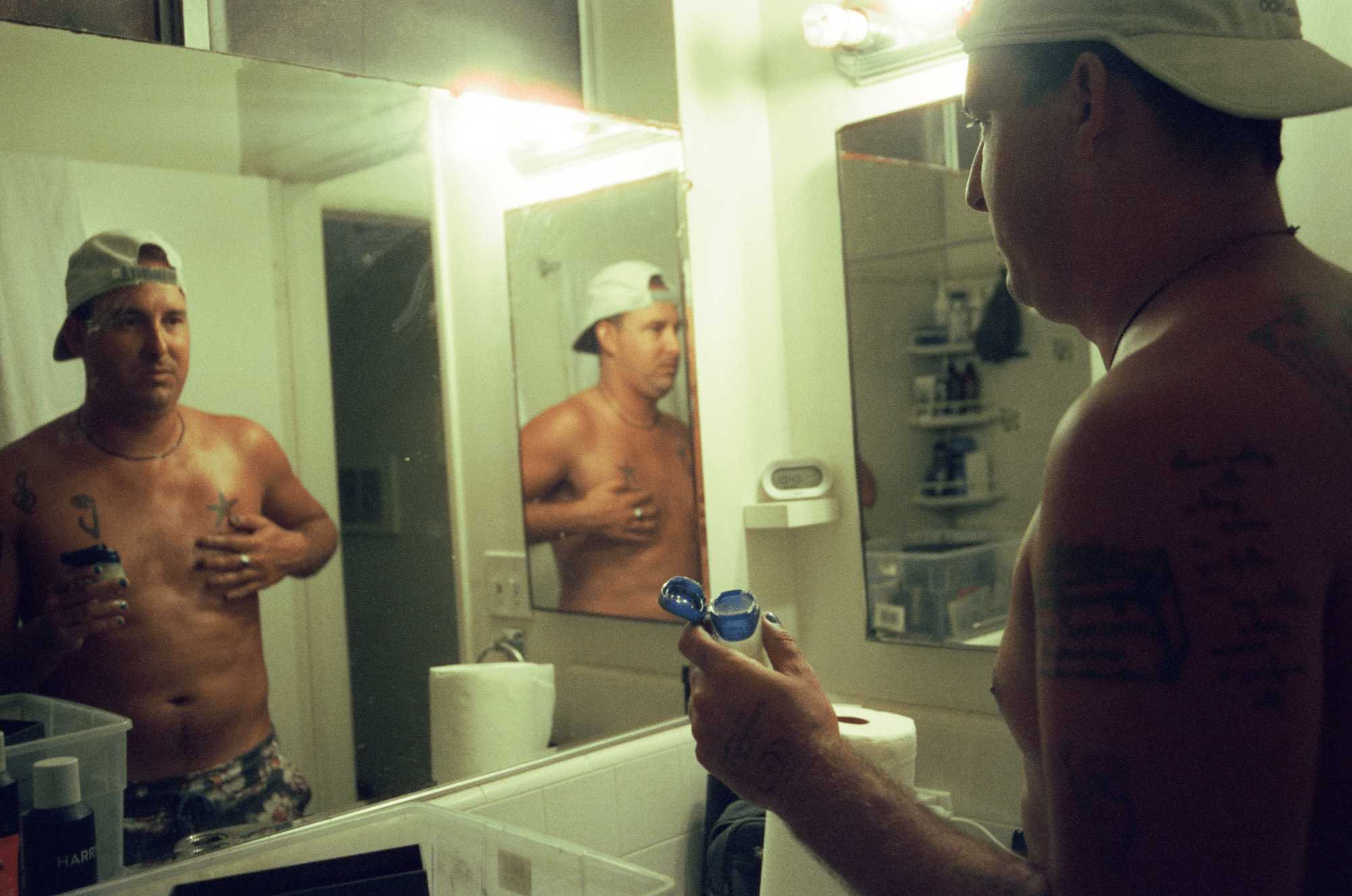 a topless man in a backwards baseball cap rubs suncream on his chest looking in the mirror