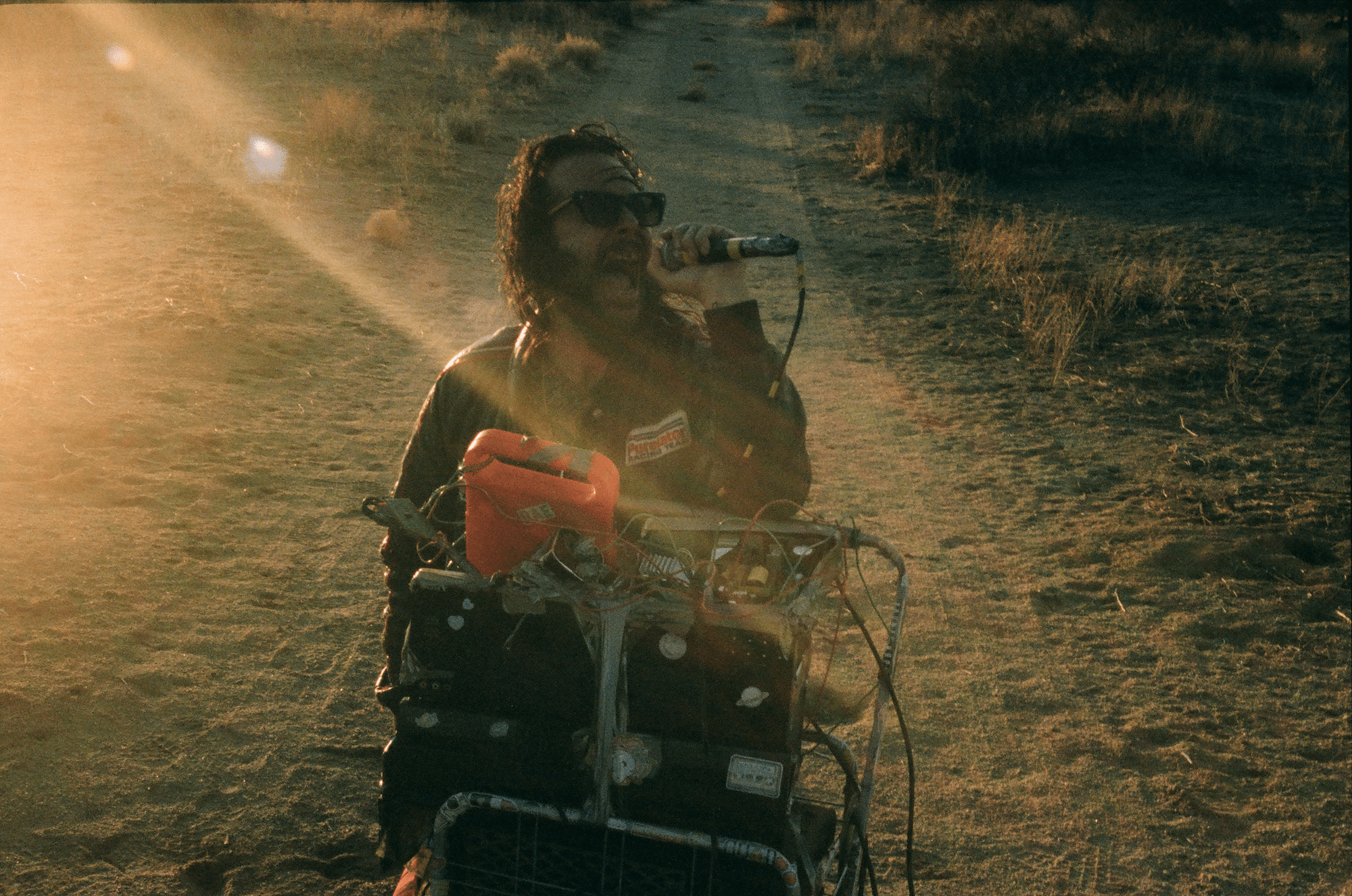 a man in a tartan shirt shouts into a microphone in a sunny desert