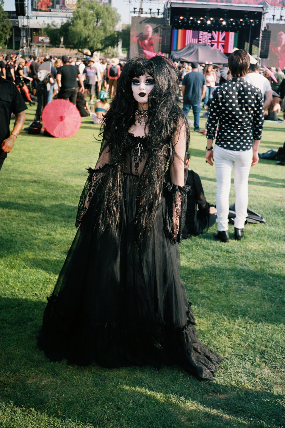 a goth girl in a long witchy black dress and a big black wig stands in front of a festival stage