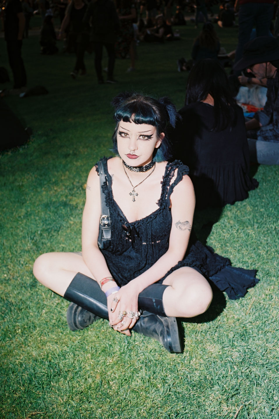 a goth girl with knee-high boots and thin painted-on eyebrows sits cross-legged on the grass