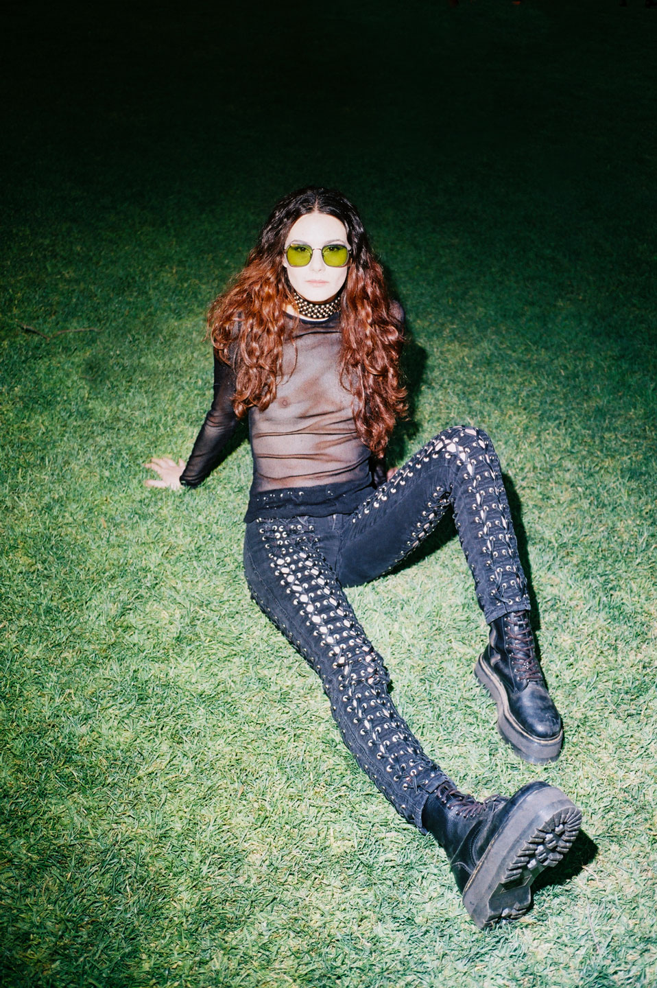 a girl wearing skinny jeans with corset detailing and a sheer black mesh top reclines in the grass