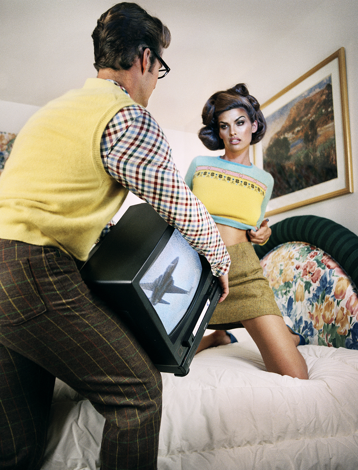 a man holding a tv in front of a model posing on a bed for a fashion editorial by guzman