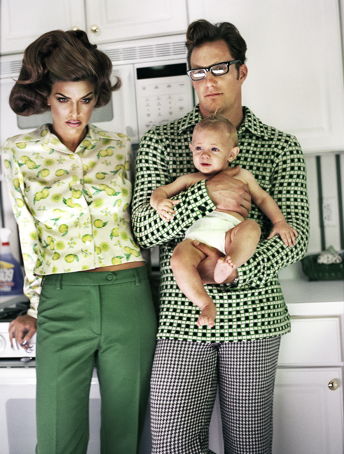 a couple posing in a kitchen in green outfits with a baby for guzman's the suburbanites