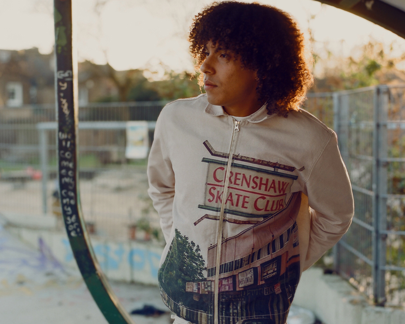 a model wears a crenshaw skate club graphic jacket, standing in a skate park