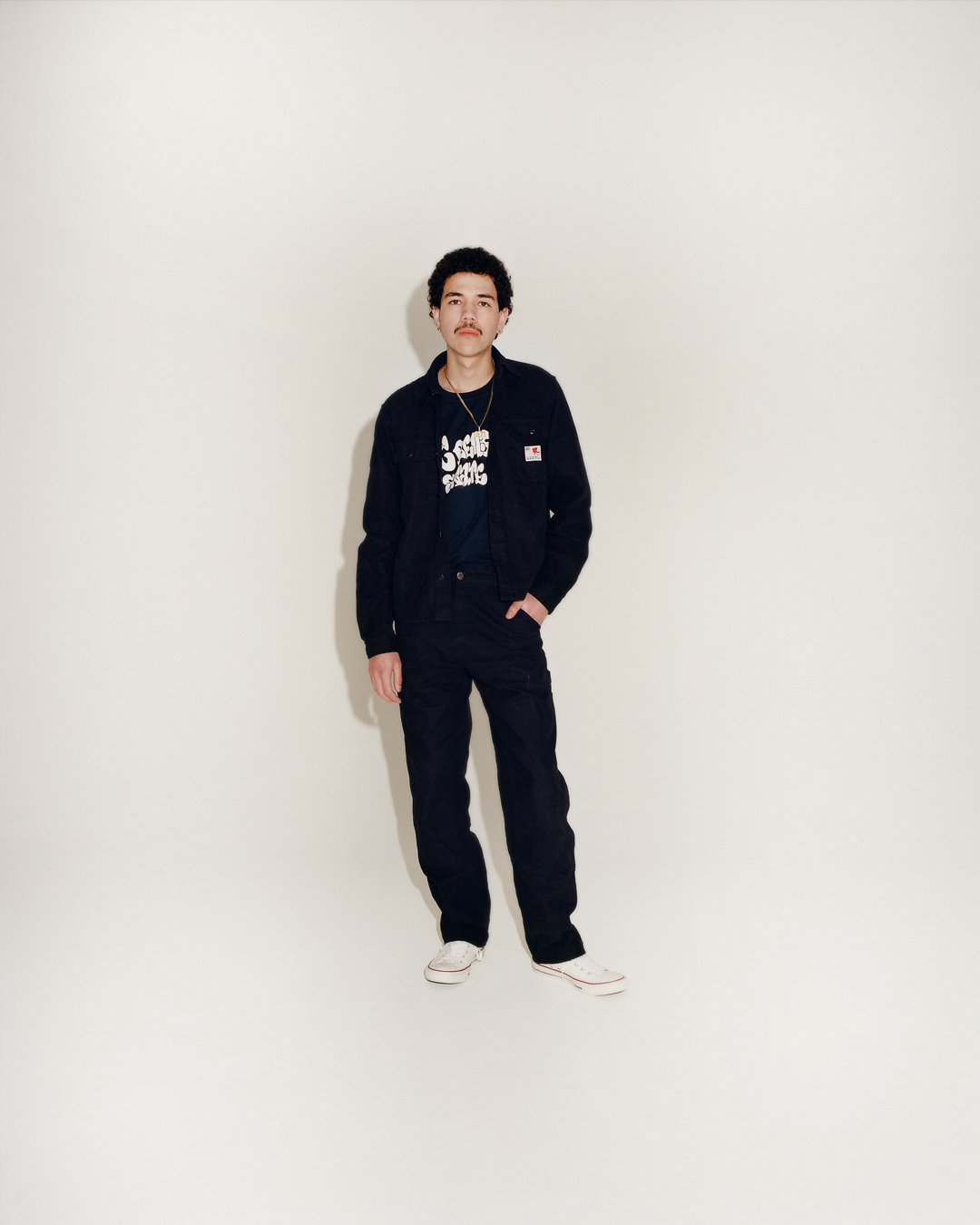 a model wearing all black stands in a white studio, hand in pocket