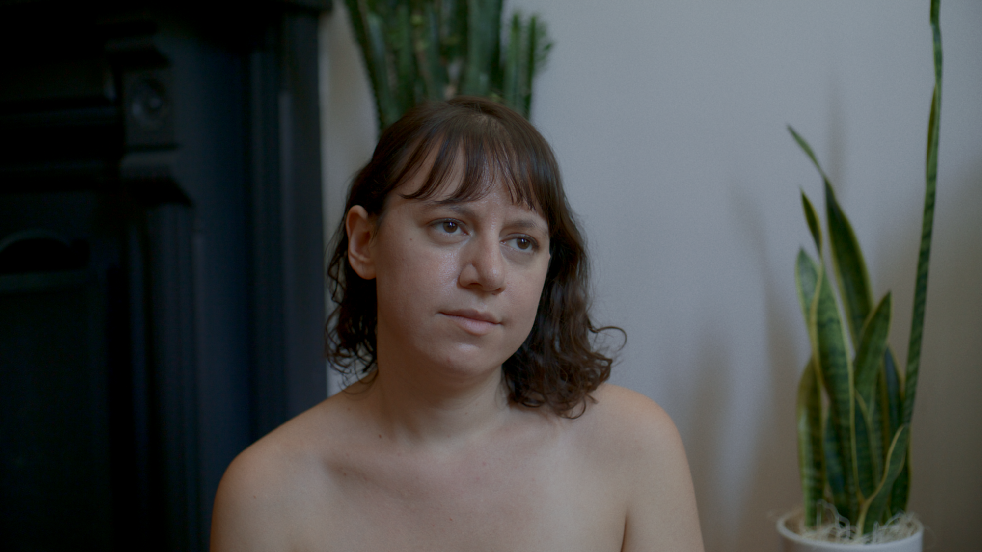 a topless joanna arnow shot in an apartment, shoulders upwards, with her curly hair back over her shoulders. two snake plants are behind her