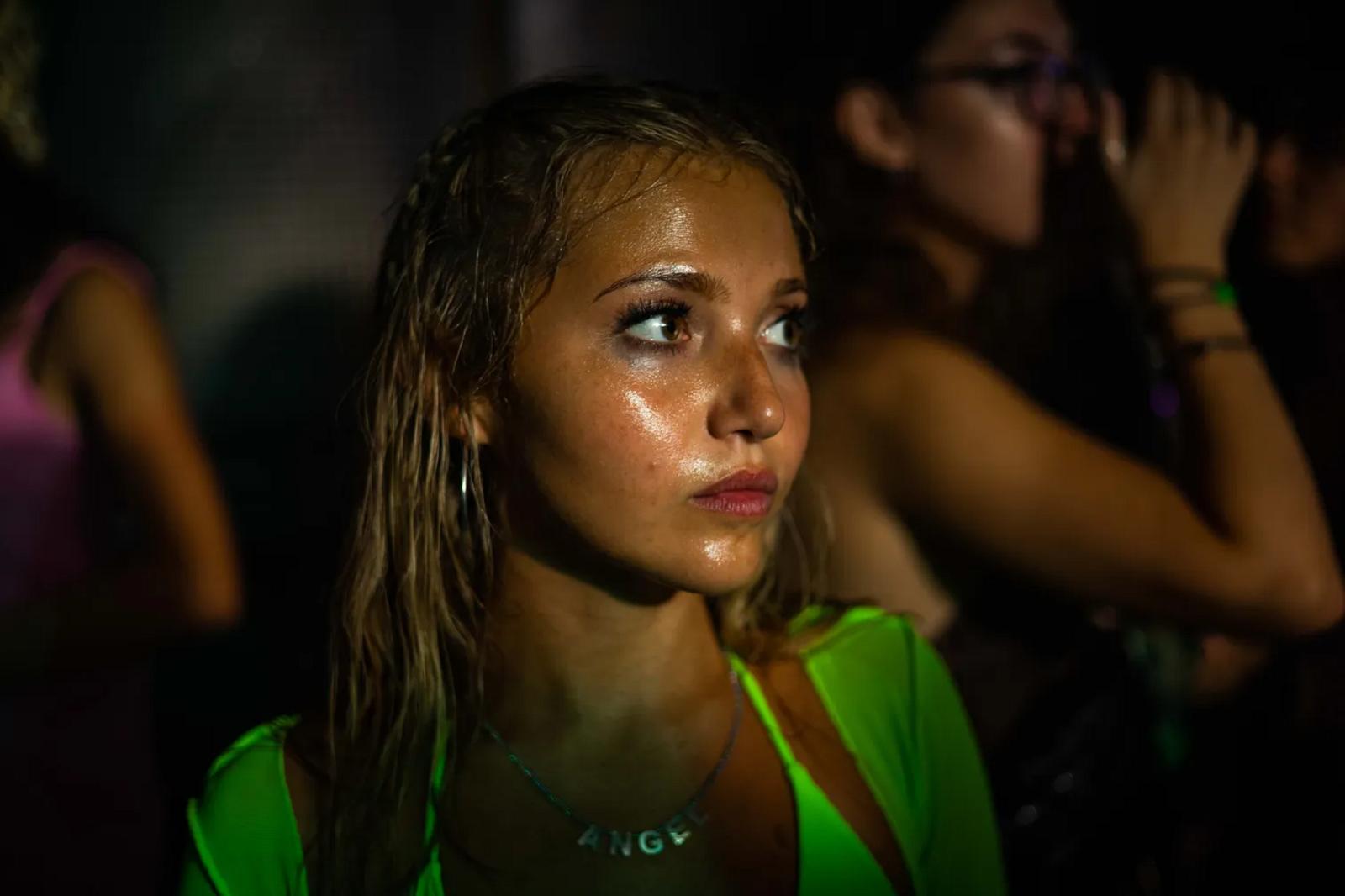 a girl wearing a neon bikini top stands in a a dark club, her sweaty face and smudged makeup illuminated