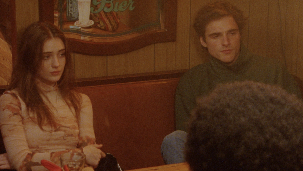 jacob elordi and talia ryder sit in a 70s looking wood-panelled bar in the sweet east