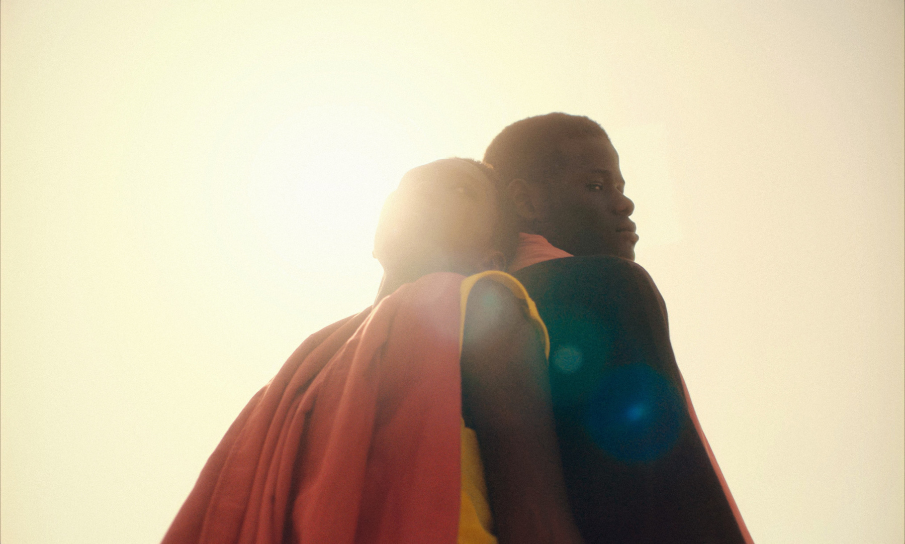 a young black couple stand back to back against the sun, shot from below