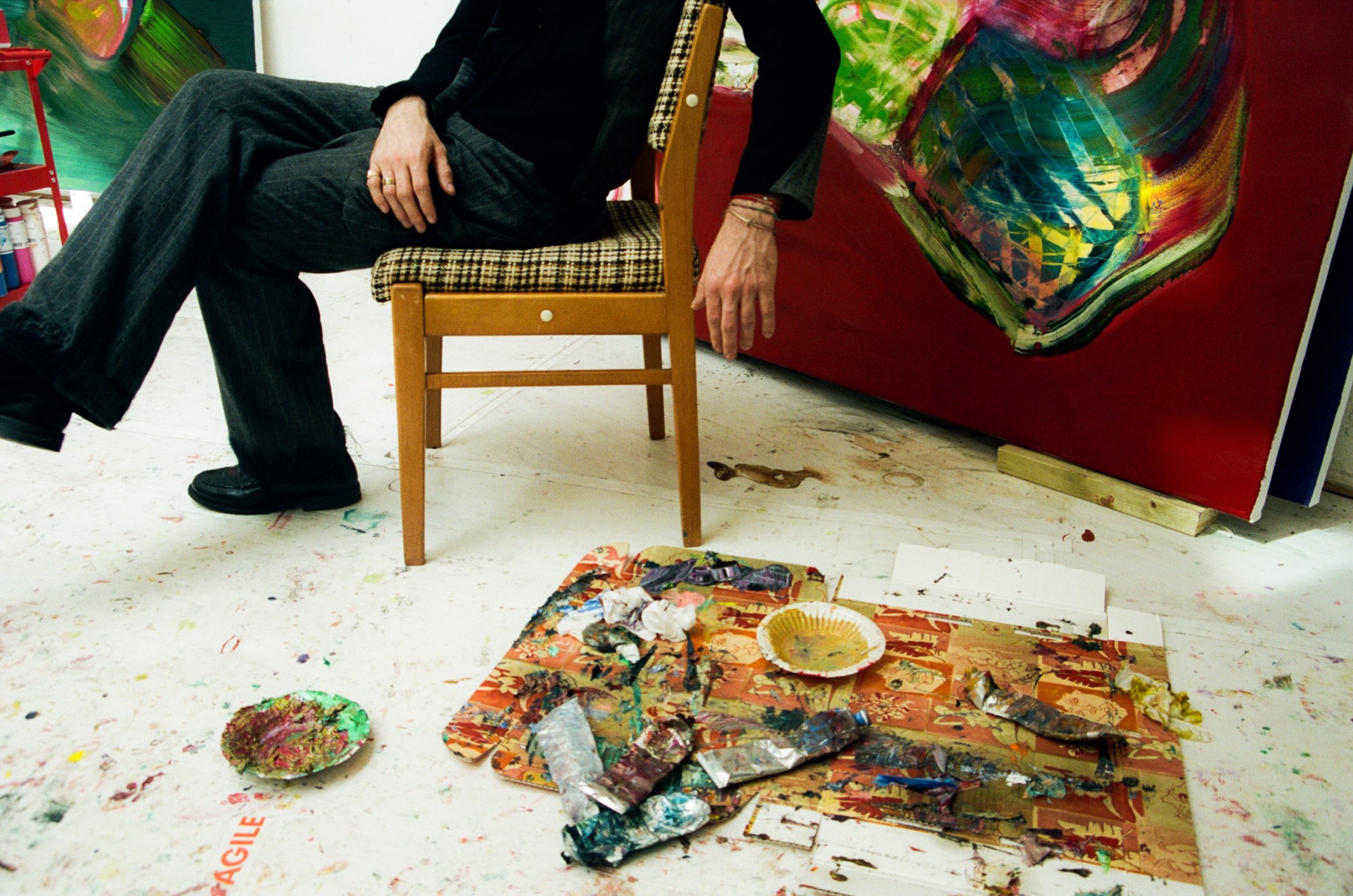 Alexander James sitting on a chair with a plaid cushion surrounded by paintings and an easel