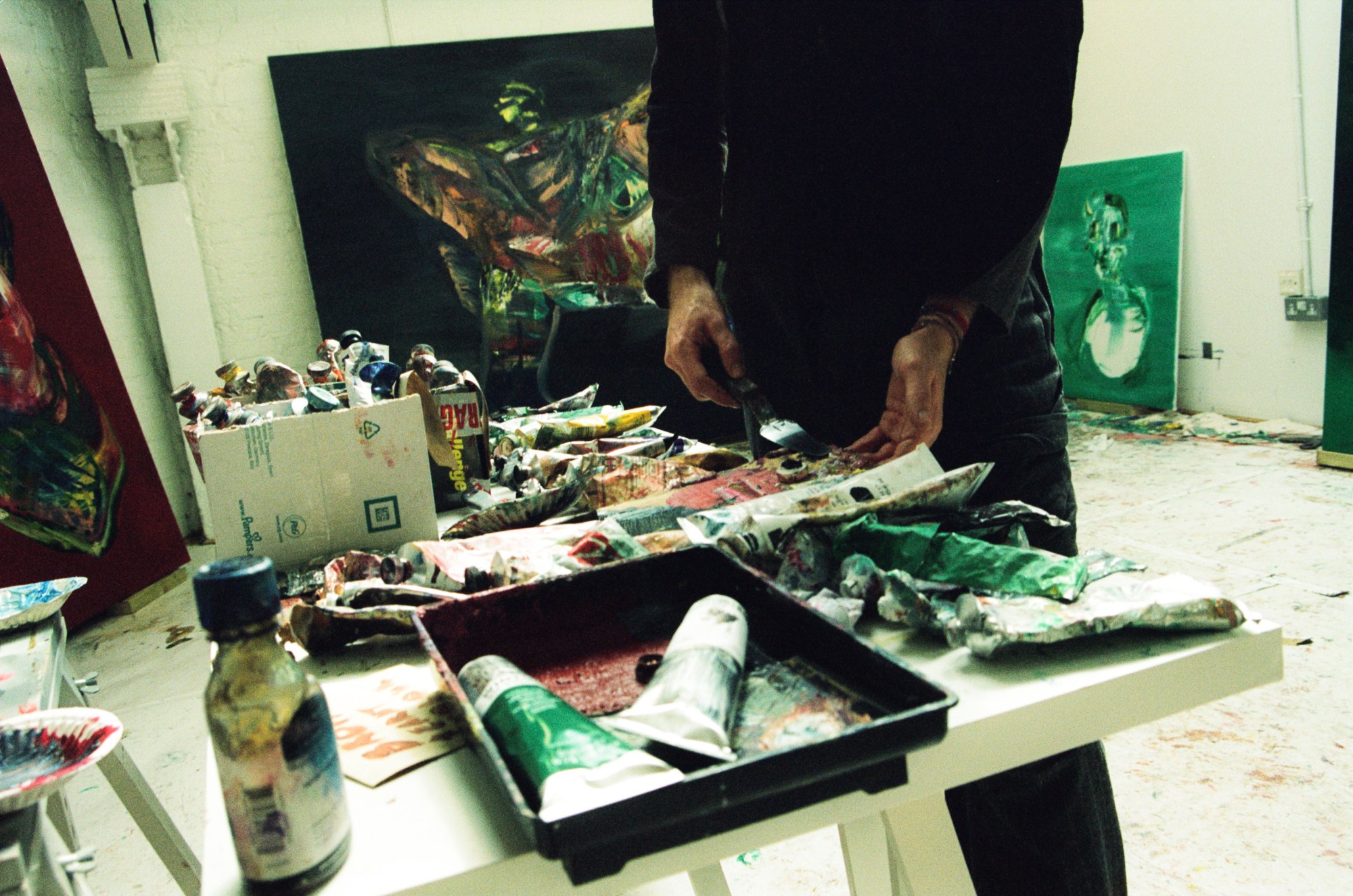 Alexander James standing in front of a table full of paint tubes with paintings in the background in his studio