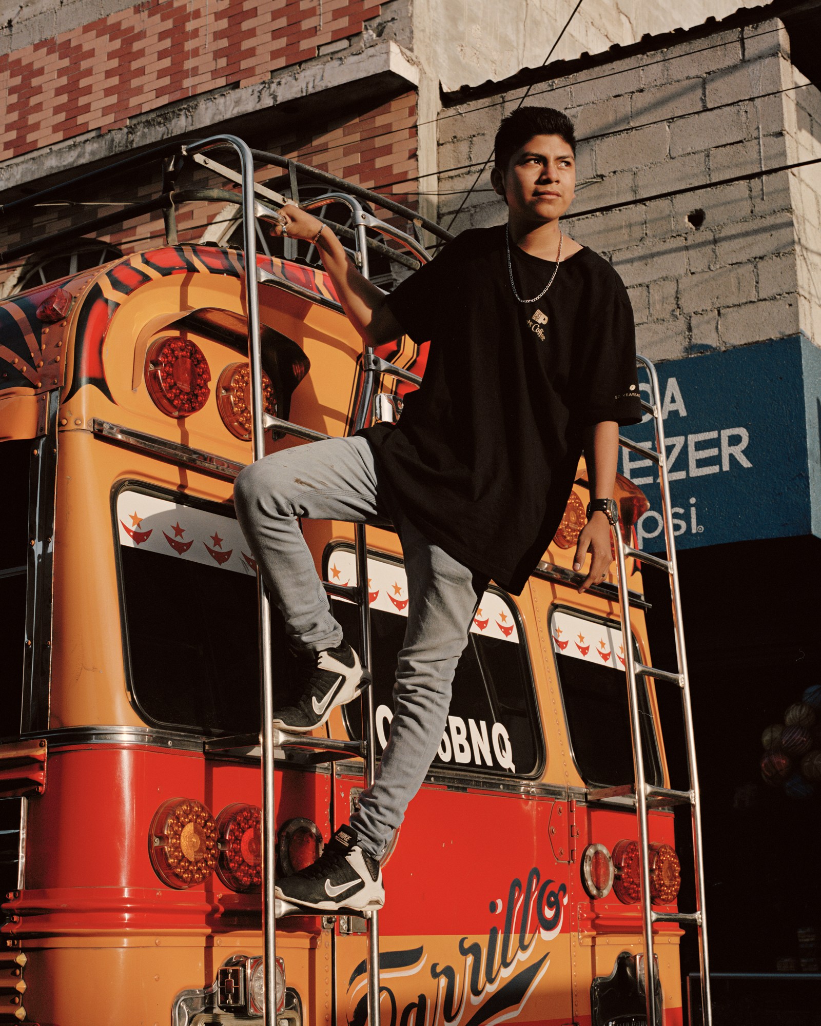 a guatemalan teen in nikes and a gold chain hanging off the back of a bus in the highlands