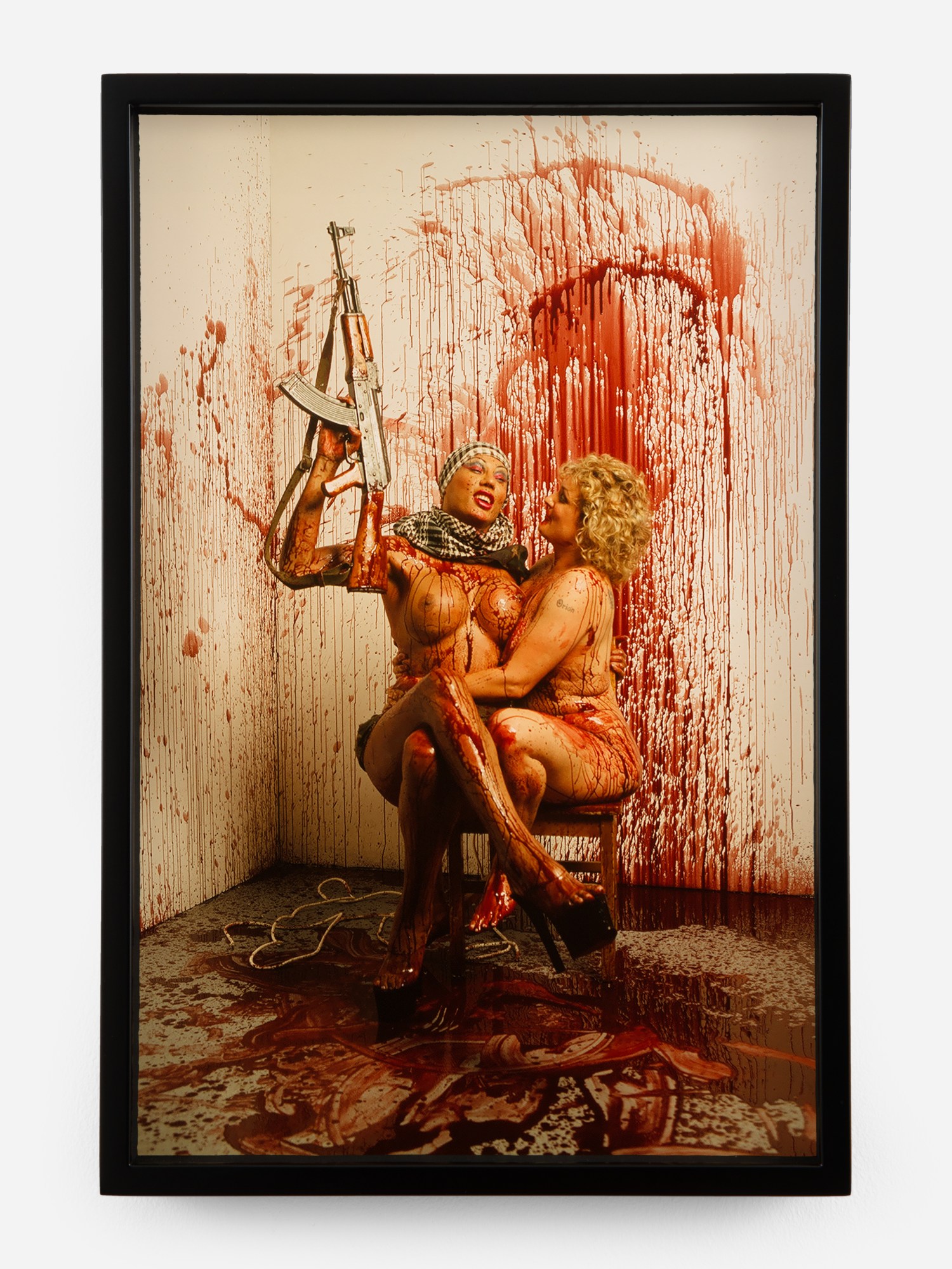 in a blood-splattered room, two nude women wearing stripper shoes sit on a chair together; one holds an AK47 above their heads