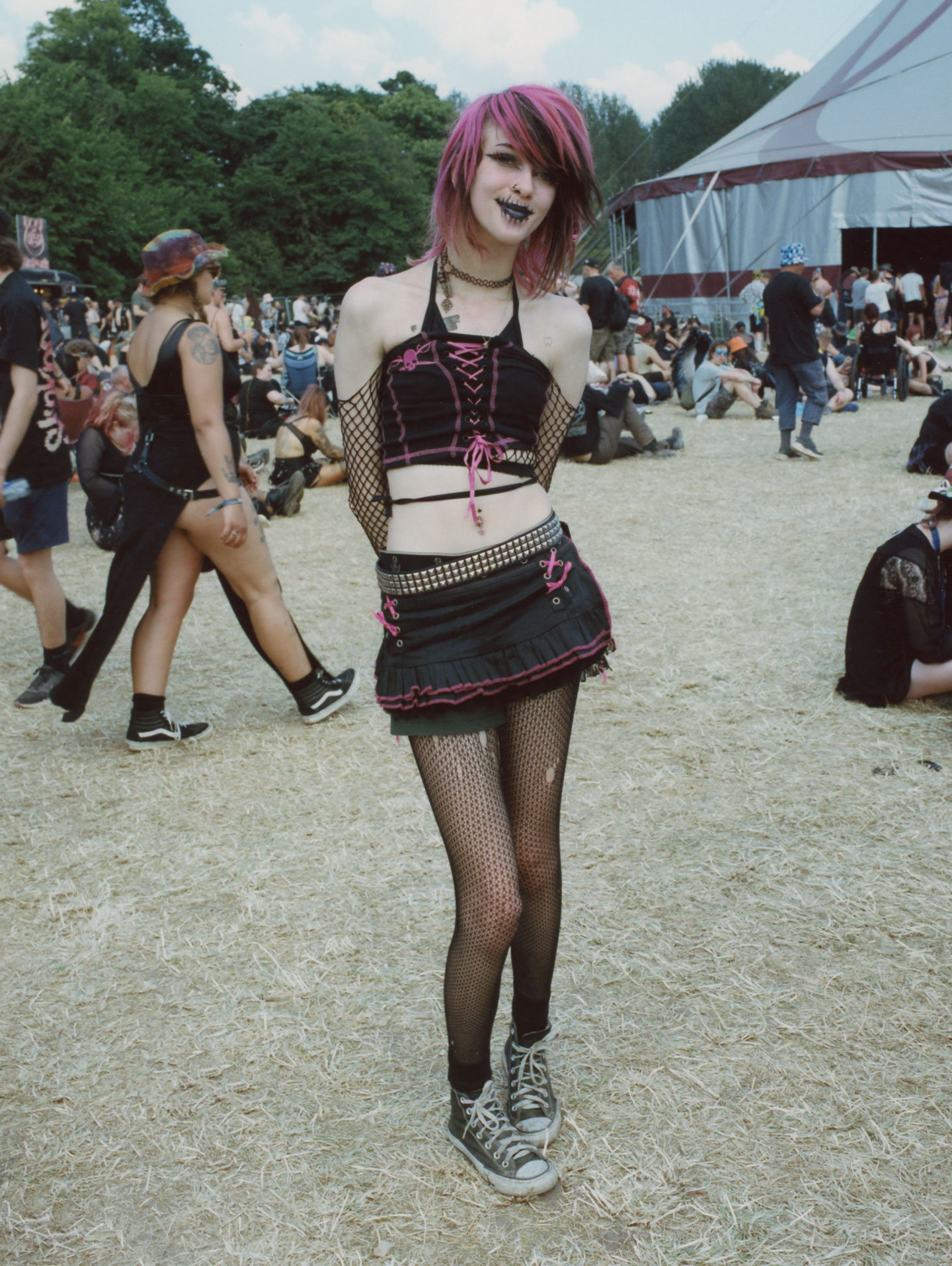 a teen with pink and black hair wears a matching corset and skirt over fishnet tights and converse; they have black lips and dark eyeliner