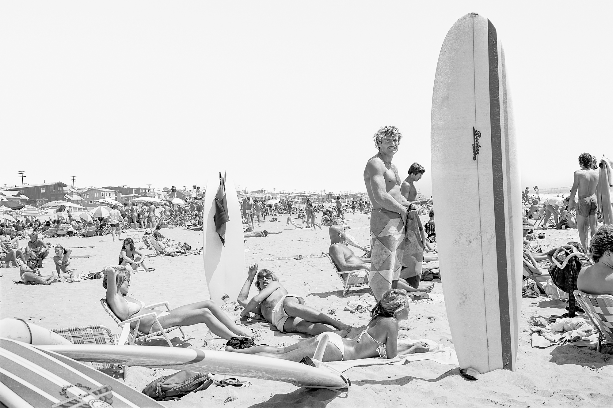 black-and-white picture of a man posing topless on a beach near a surfboard