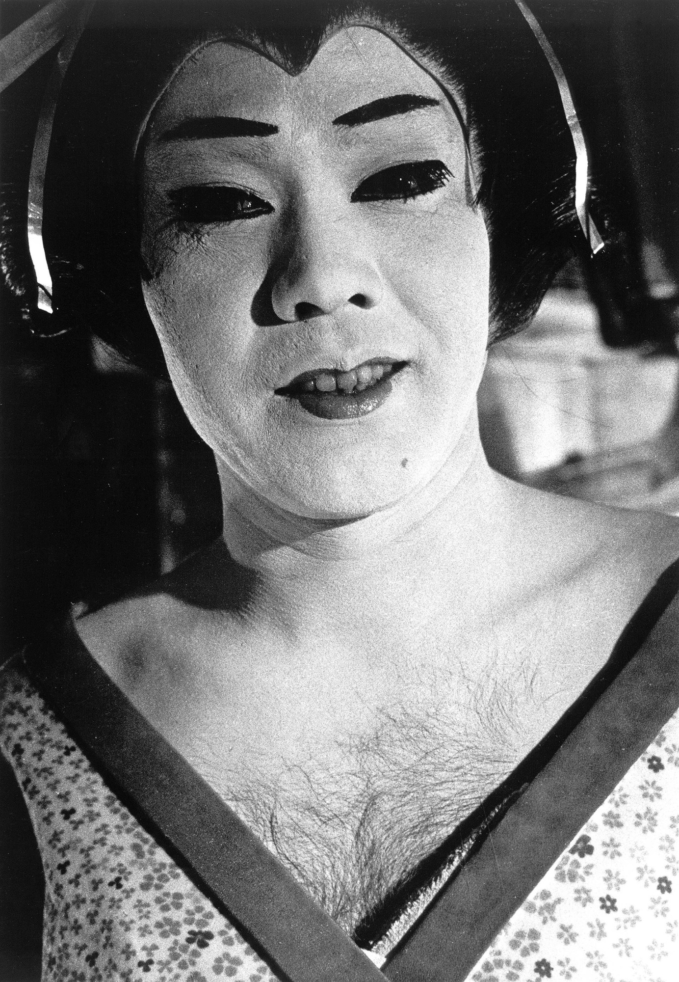 a black-and-white photo of a person wearing geisha make-up and a wig. their teeth are showing. they have a hairy chest and are wearing a floral kimono.