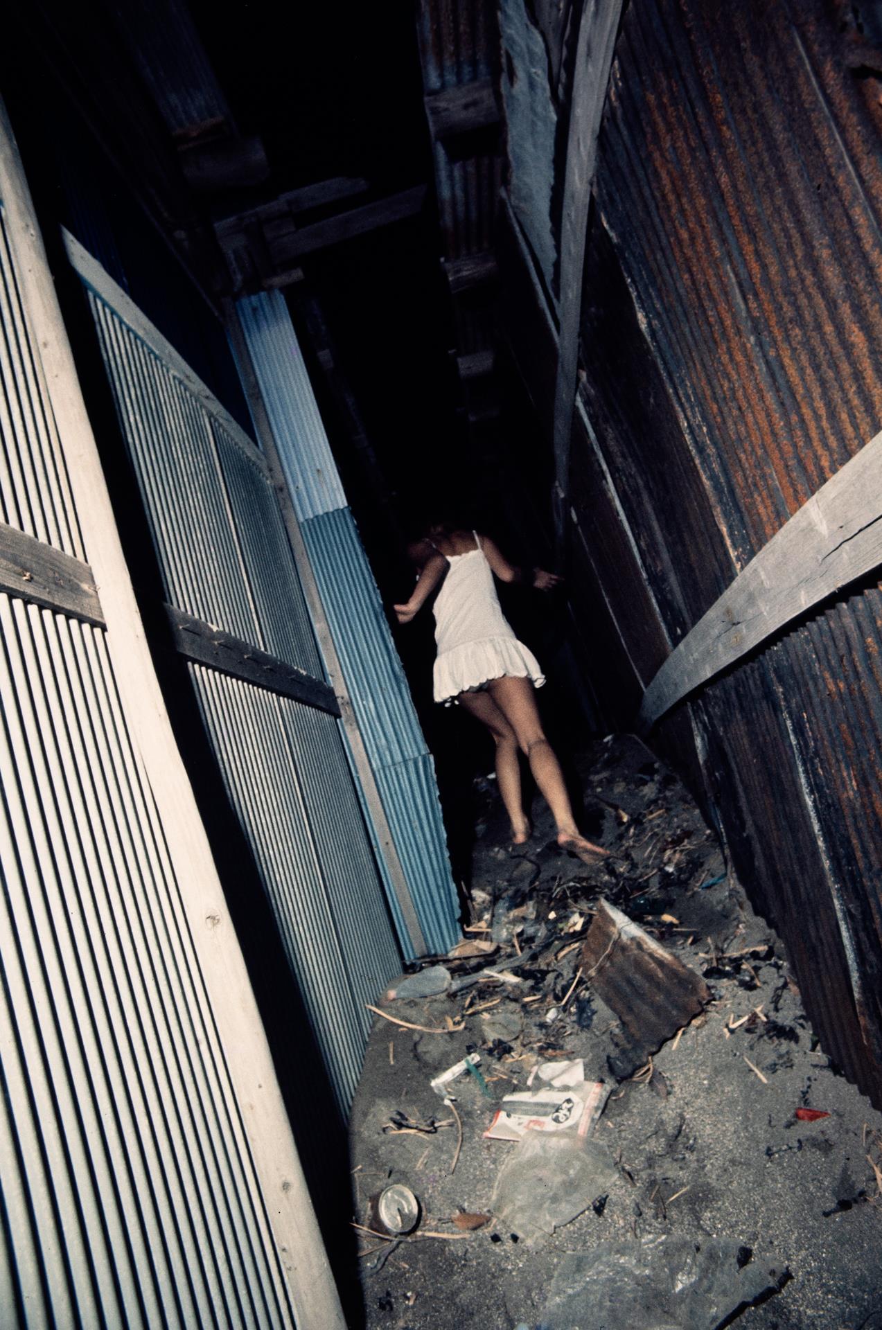 a woman in a white dress runs up a dirty alleyway covered in rubbish away from the camera.