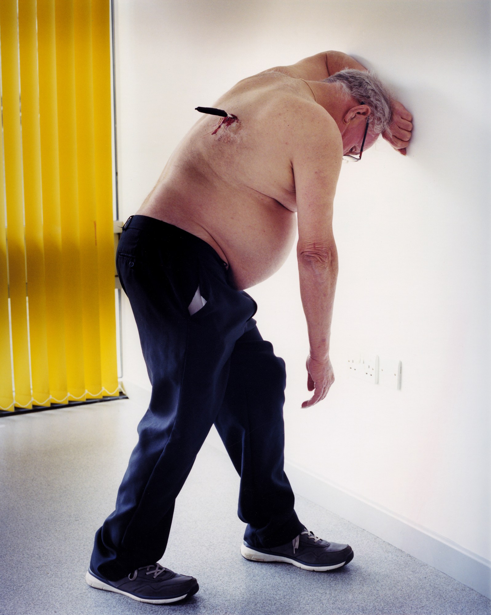 a portrait of a topless man in sneakers and jeans lying against a wall with a fake knife in his back