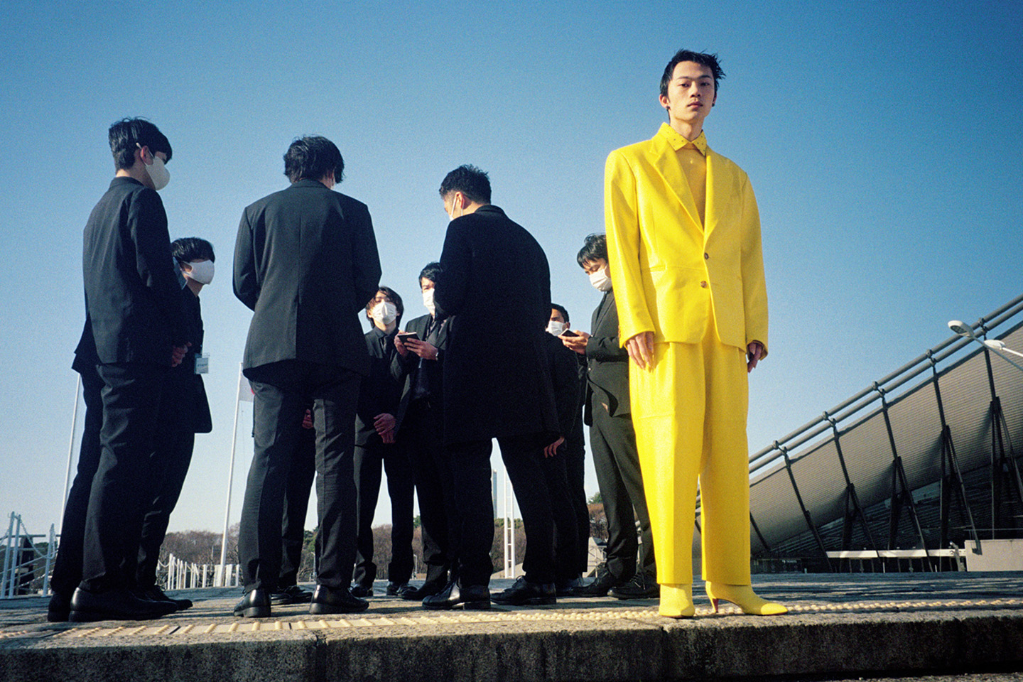 a model in a bright yellow suit (and yellow heeled boots) stands on a concrete platform next to a group of men in black suits wearing face masks