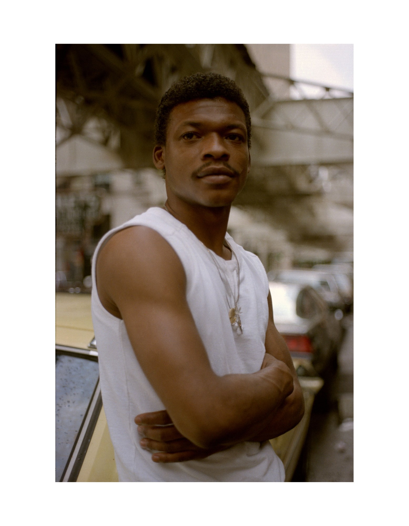 a man in a white tank top and gold necklace posing for a portrait while leaning on a yellow car