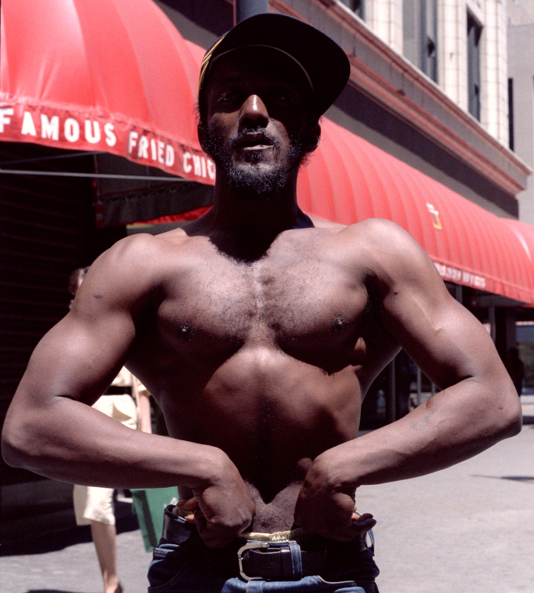 a man flexing for a potrait wearing belted jeans and a baseball cap