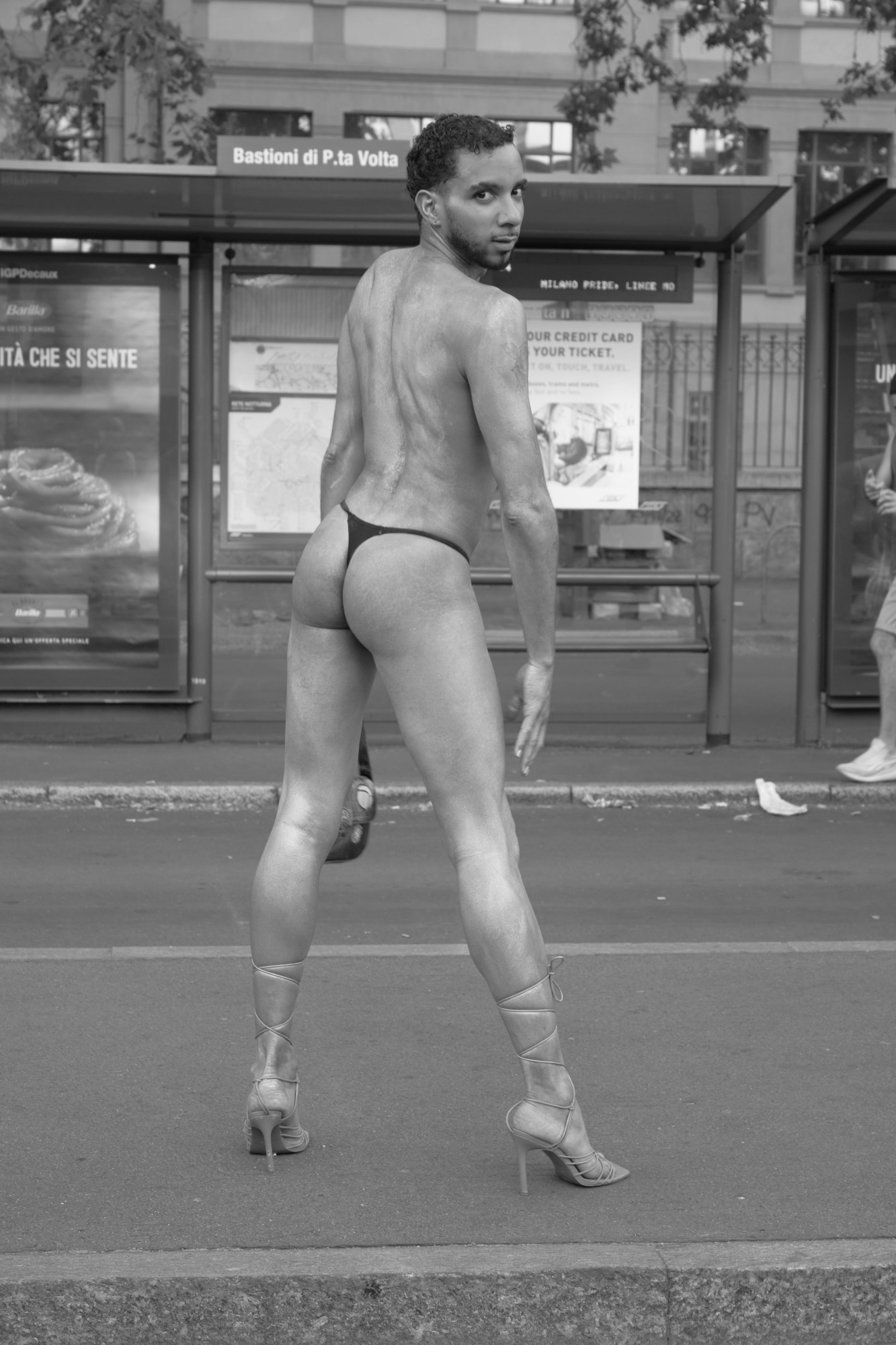 a person at milan pride stands in front of a bus stop looking over their shoulder. they are wearing a thong and strap up stiletto heels.