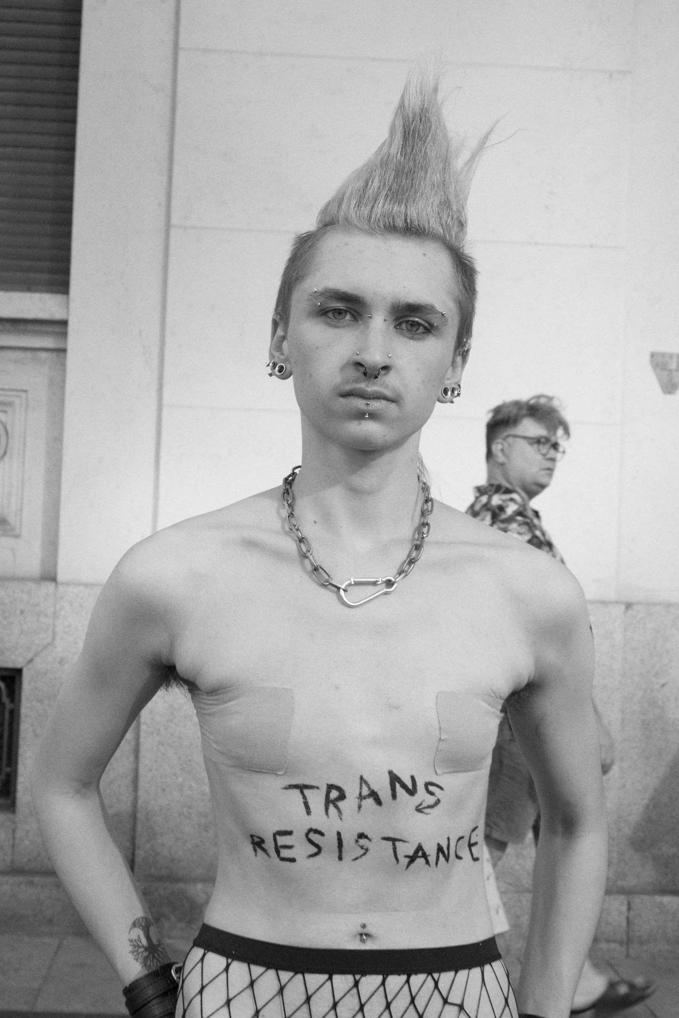 a transmasc person is topless wearing binders and fishnet tights. their hair is in a mohawk and the phrase 'trans resistance' is written across their stomach.