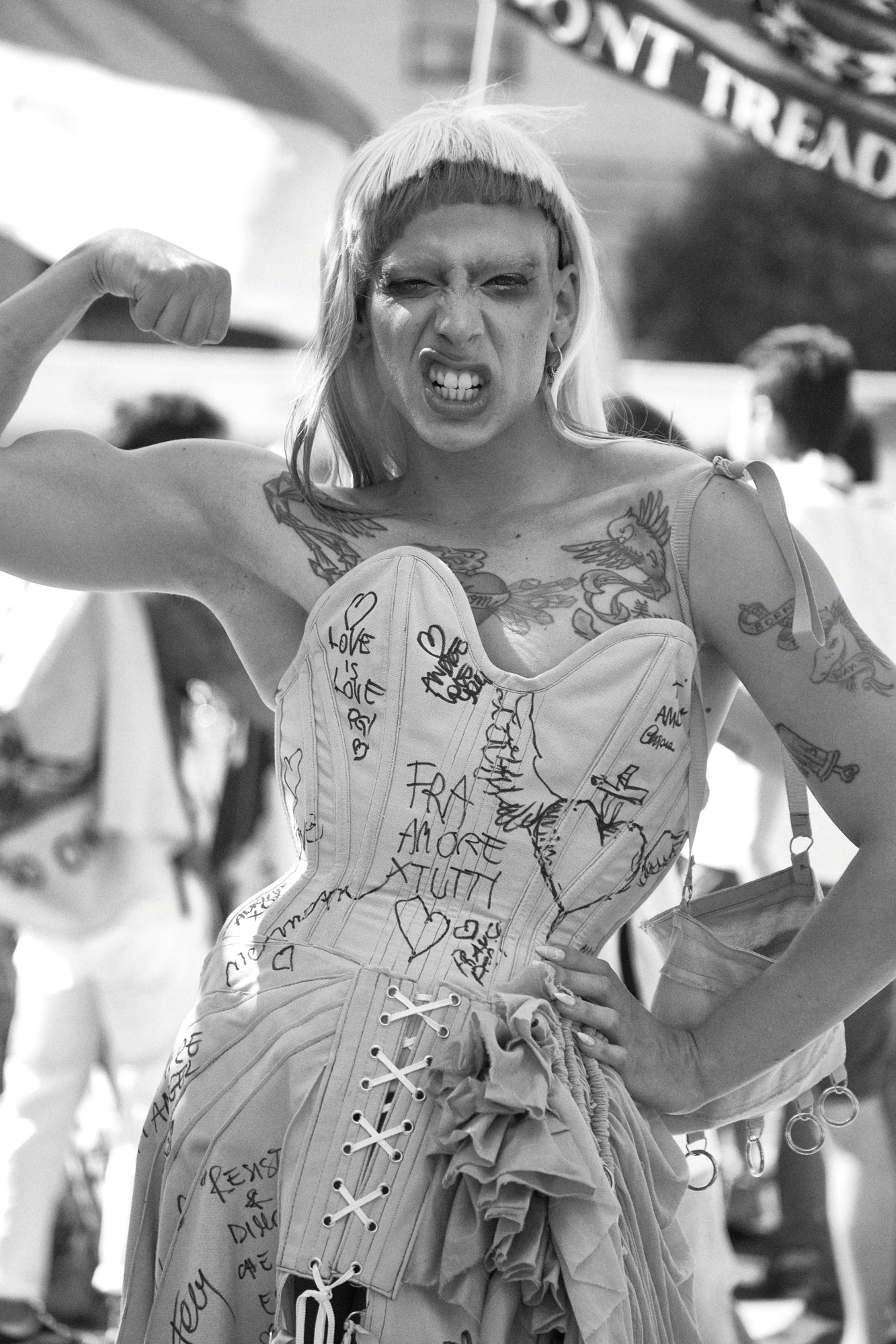 a person wearing a light corseted dress covered in phrases like resist and love is love poses for the camera by flexing their bicep at milan pride.