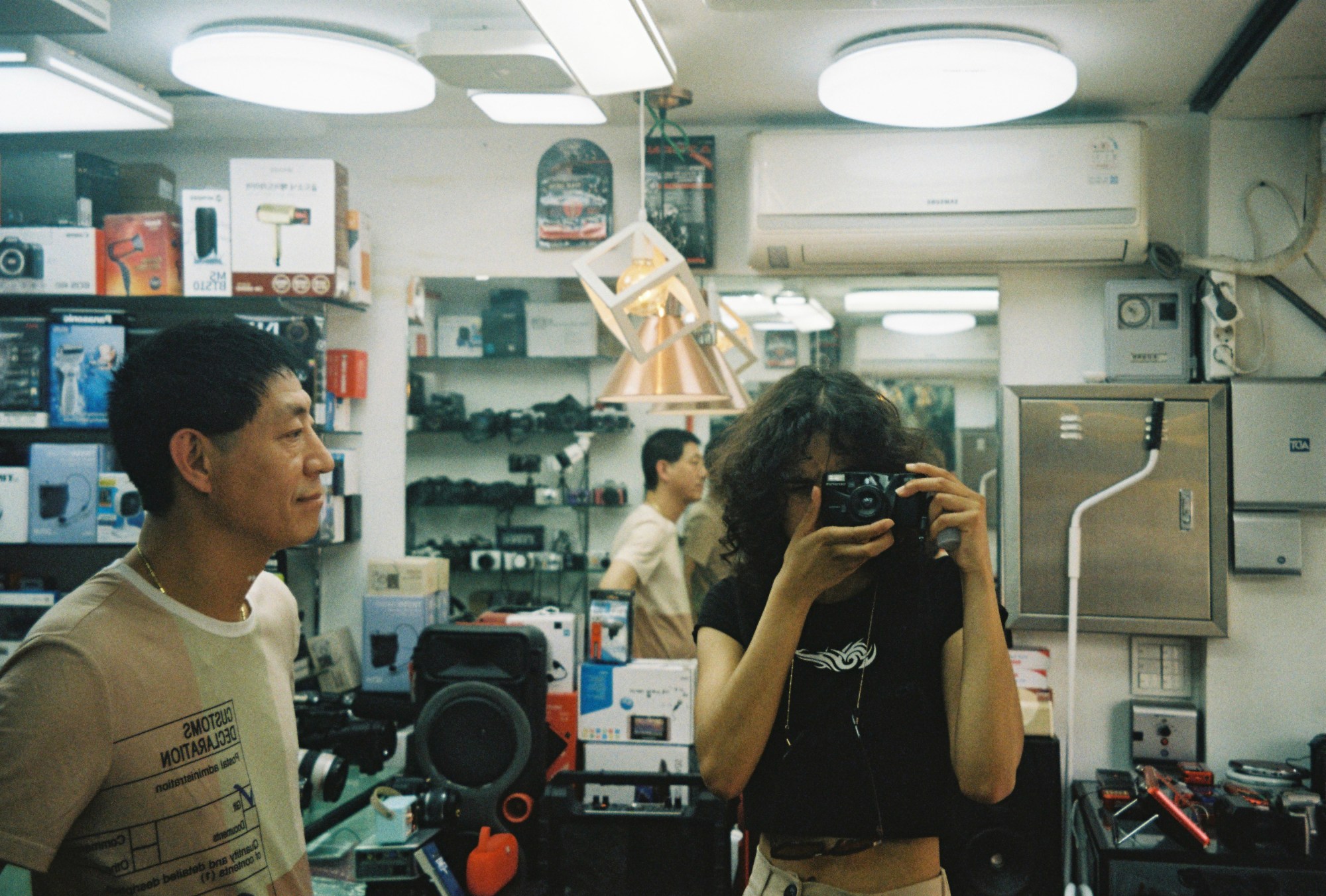 a man watches a woman taking a picture of herself with an analog camera