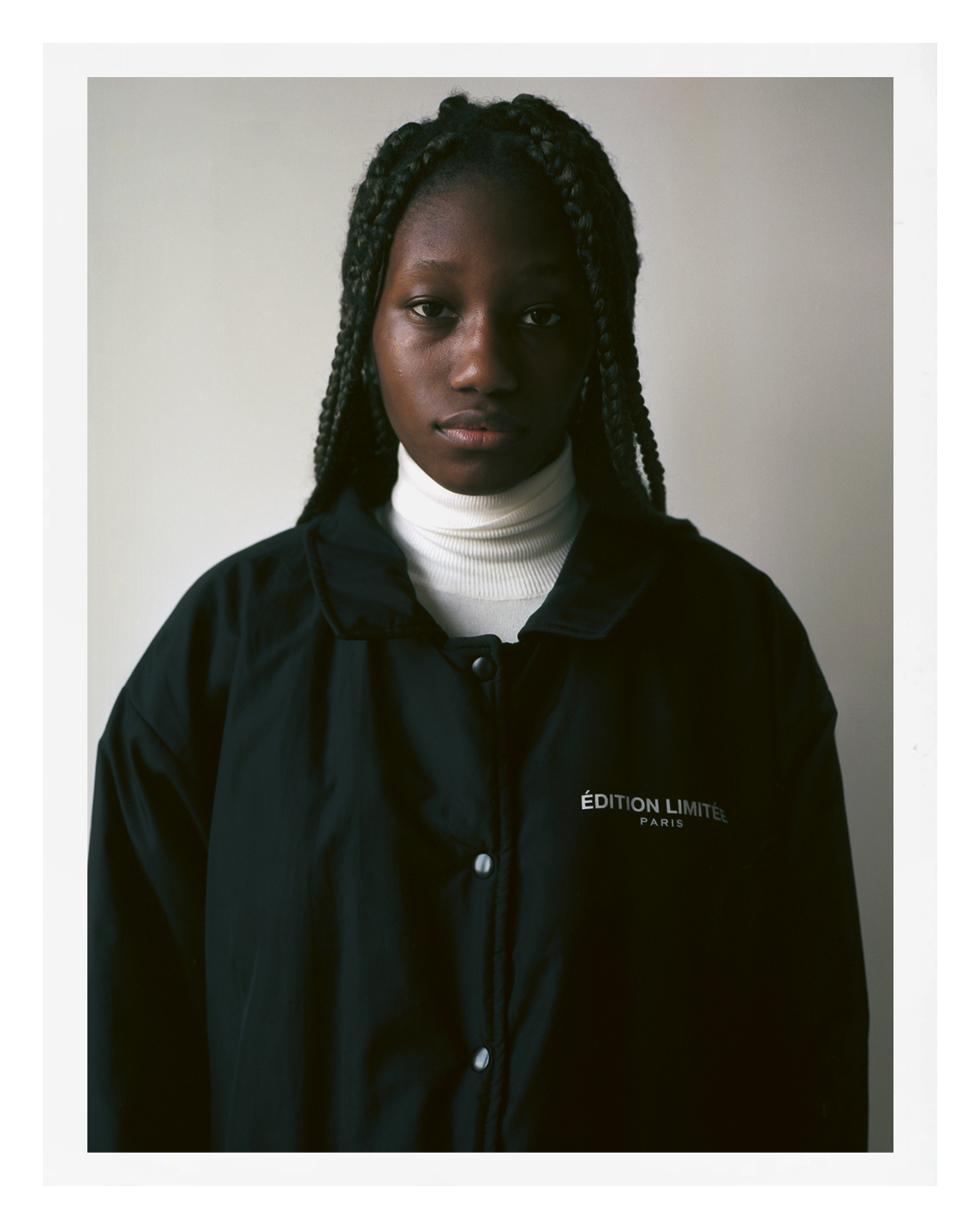 photo of Black parisian teenager standing against a white wall wearing a black jacket and white rollneck