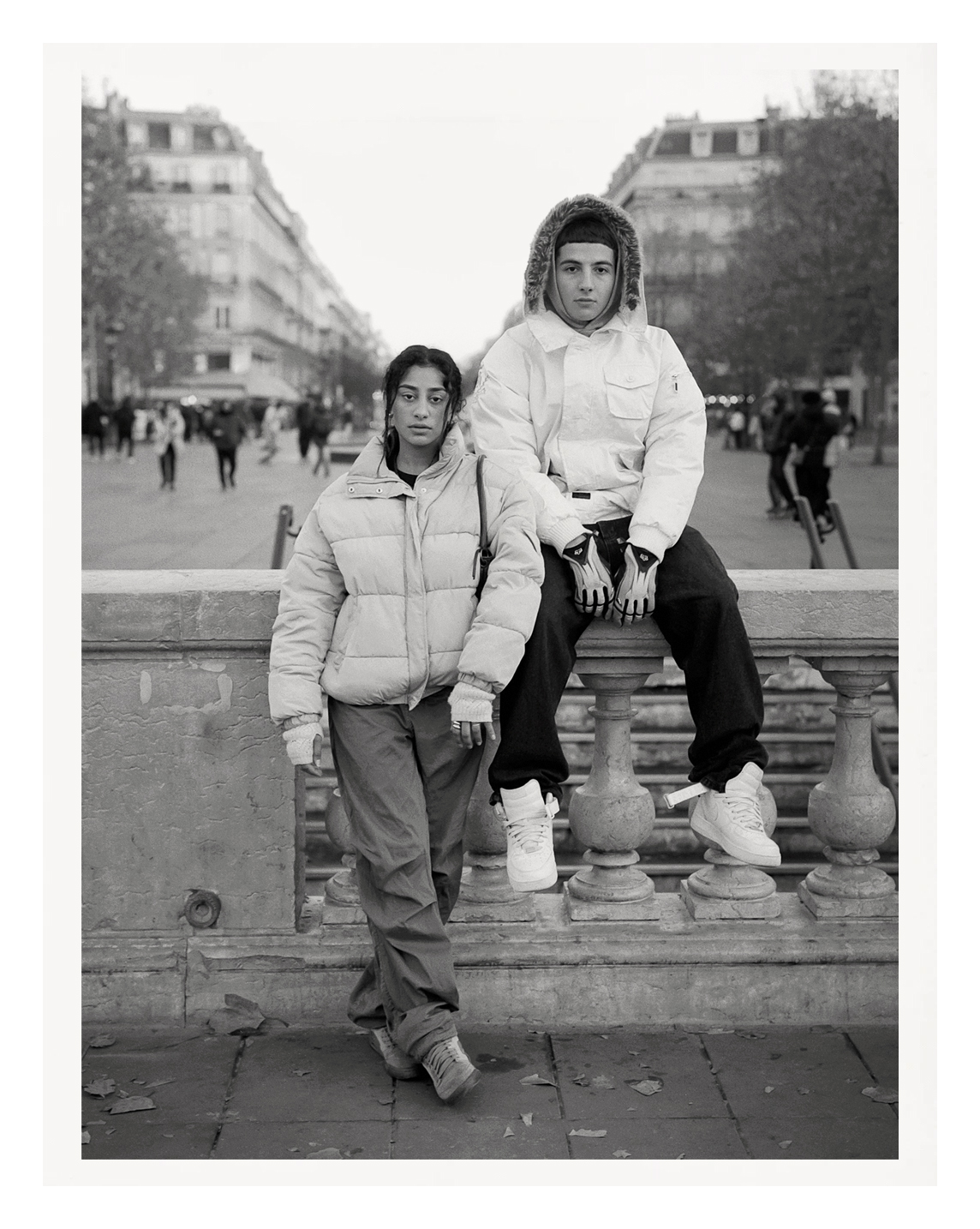 two young parisian siblings wearing heavy jackets look into the camera lens, perched on a stone wall.