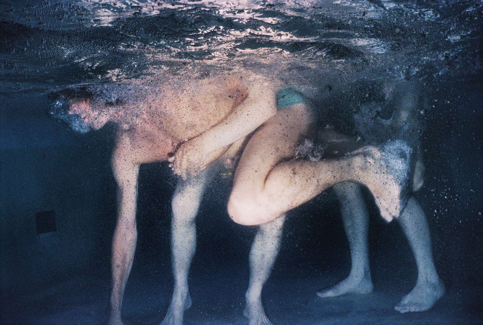 an underwater shot of a man reaching down to touch the bottom of a pool; the legs of two other swimmers are seen standing behind him