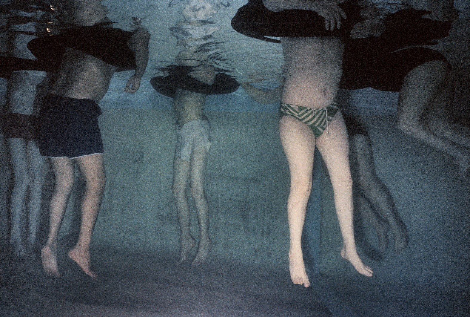 an underwater shot of six people floating in a swimming pool, rubber rings around the tops of their bodies for support