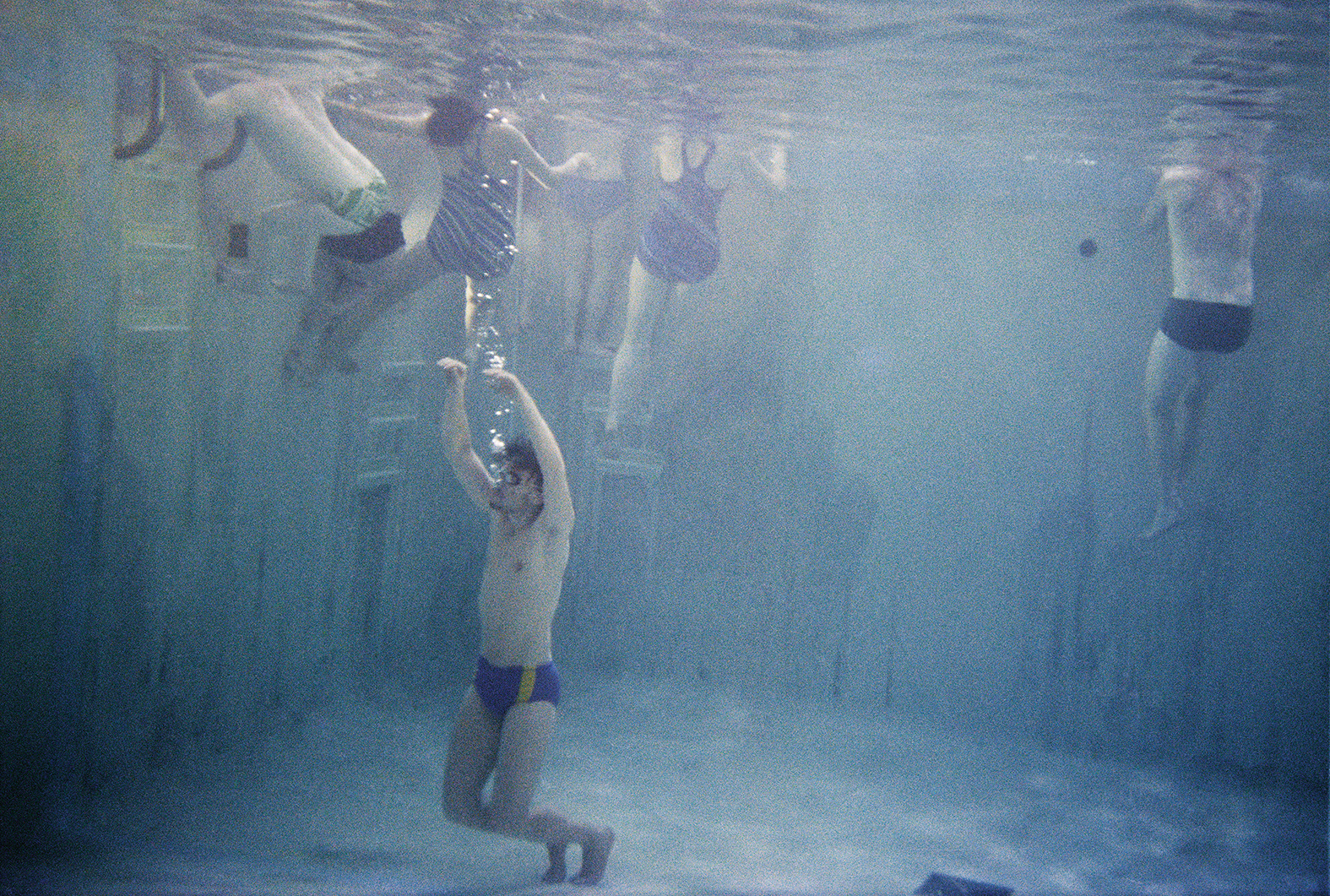 an underwater shot of six people in a swimming pool; one of them is at the bottom of the pool, arms in the air, sending bubbles up to the surface