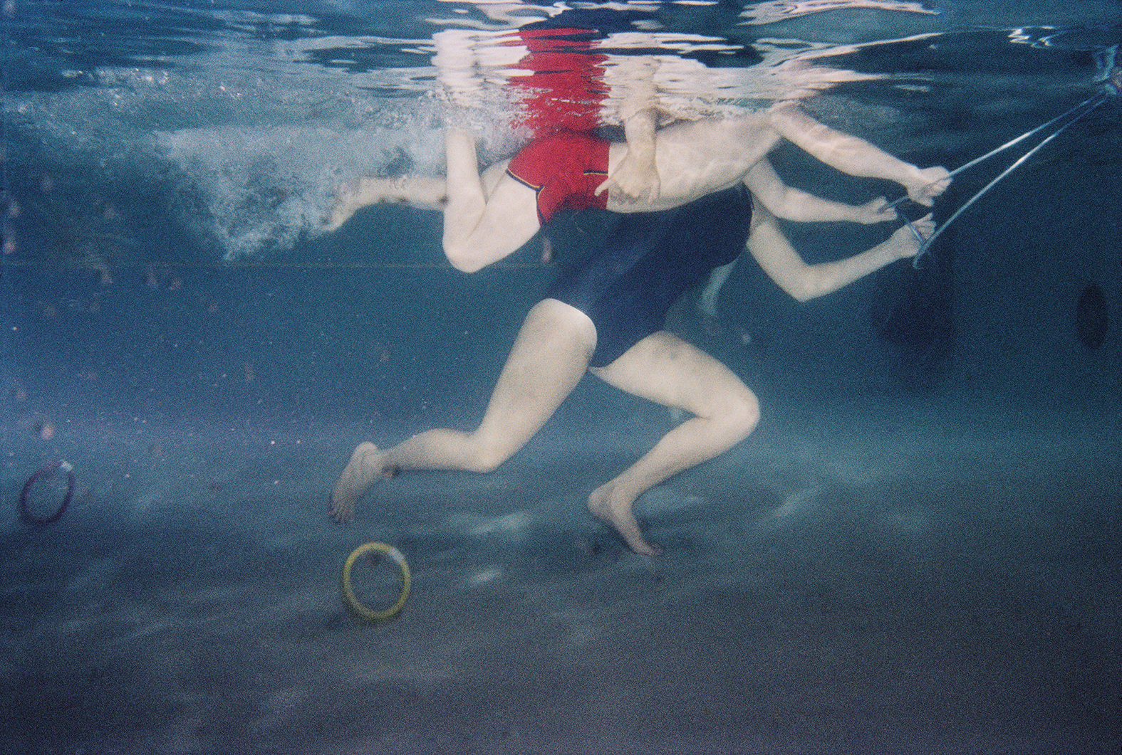 an underwater shot of two people in a swimming pool learning to swim; they hold onto a rope presumably being pulled along; there are colourful rings laying on the floor