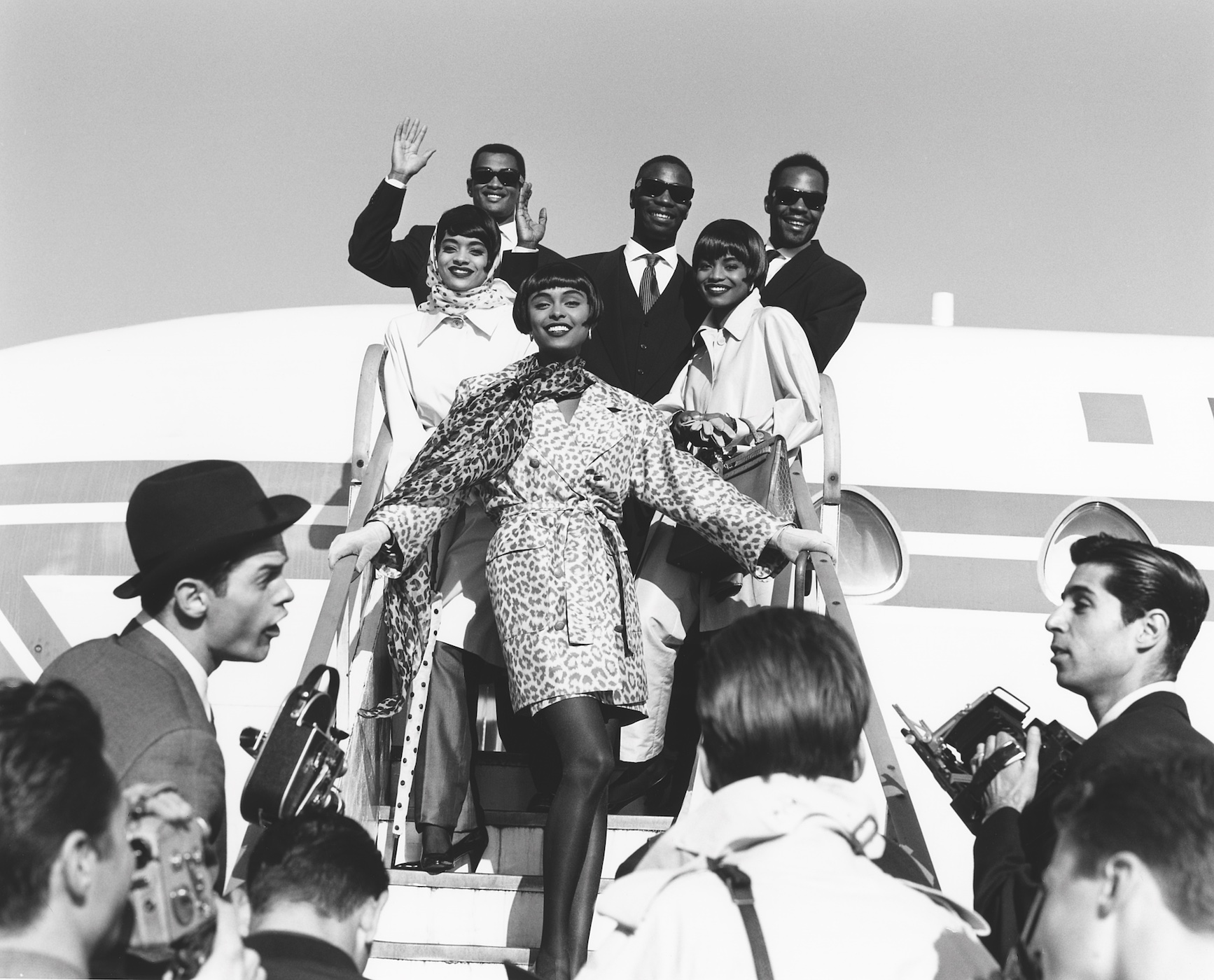 black-and-white image of models descending from an airplane as journalists wait on the steps