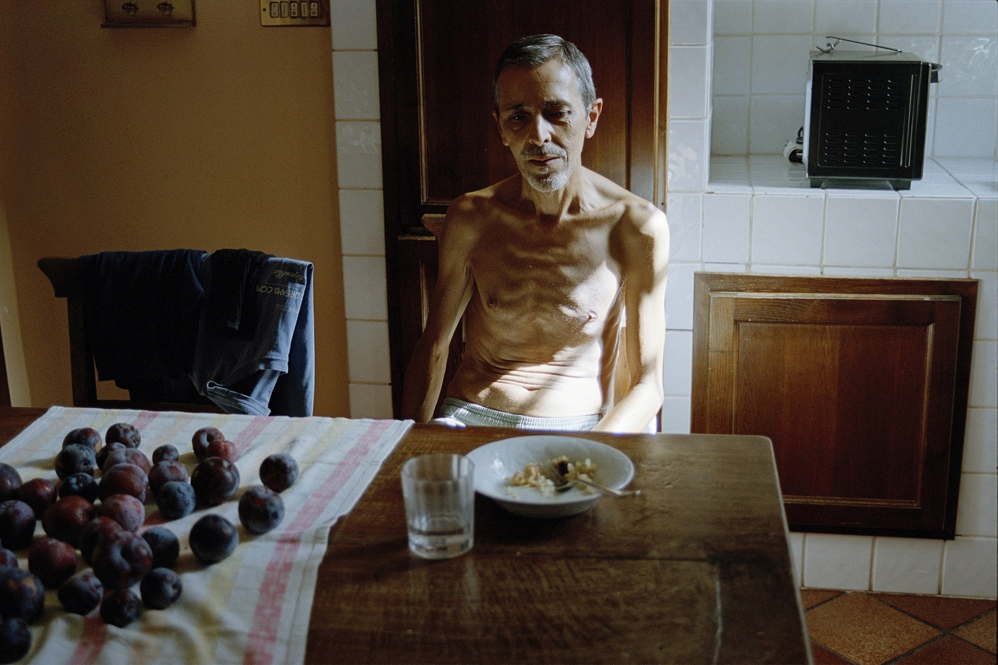 a topless man sits in a kitchen at a table with a glass, a bowl and a selection of plums on a tea-towel