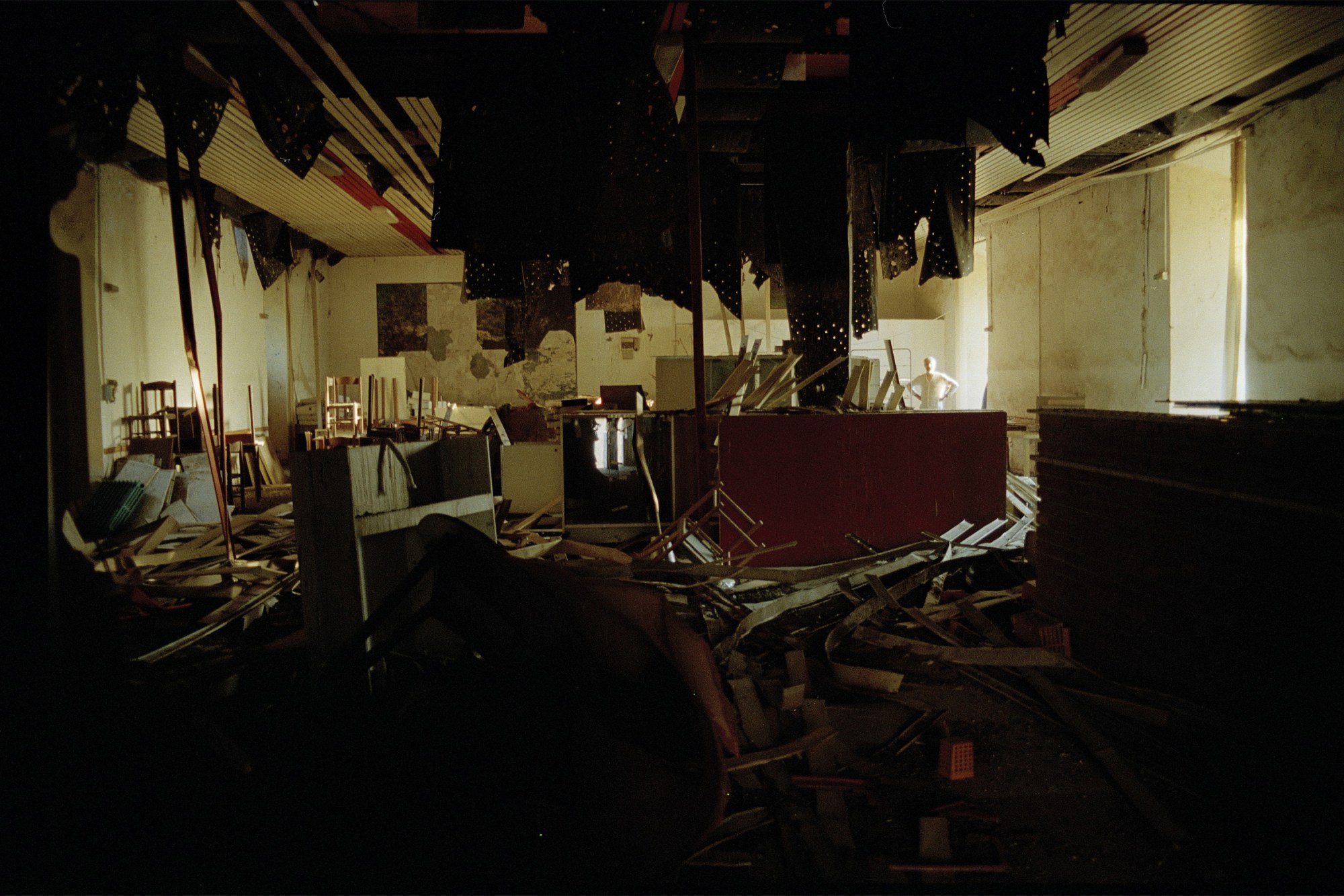 an abandoned and decaying building full of wreckage from a collapsed ceiling, in the far back background a person surveys the room