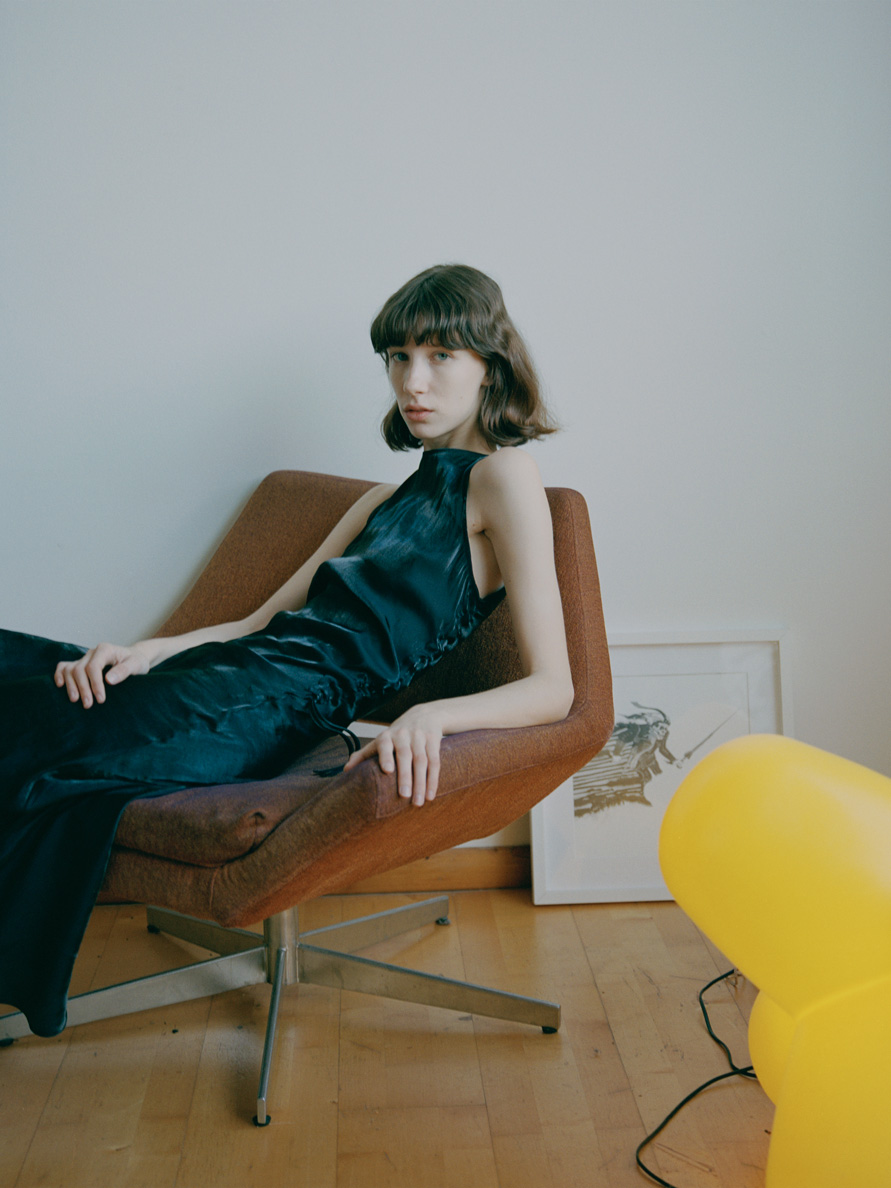 a white model with a brown bob reclines casually in a mid-century chair; an abstract yellow light stands in the foreground