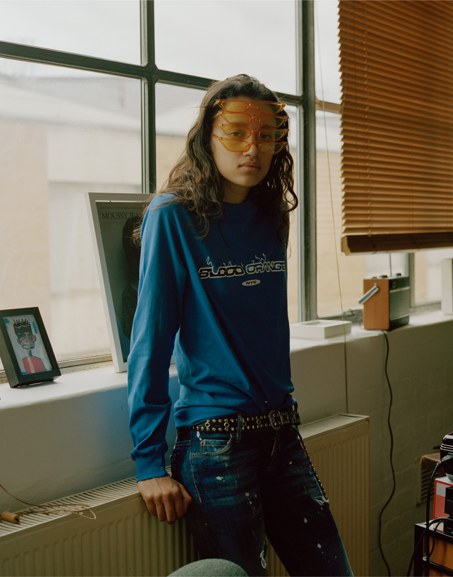 a brown model wearing blood orange merch balances four pairs of comedy sunglasses on her face as she leans against a window