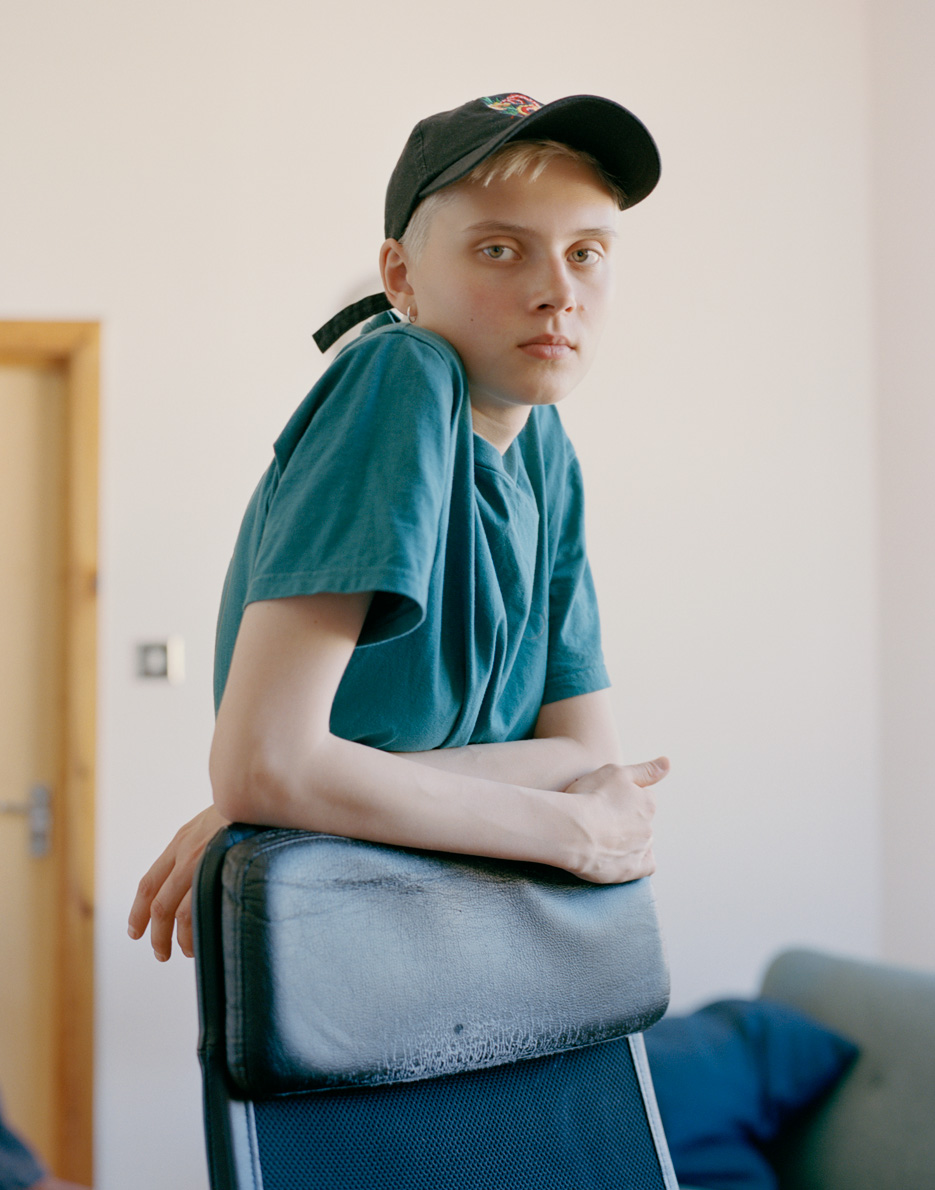 an androgynous blonde model in a blue t-shirt and cap leans against a mid-century chair