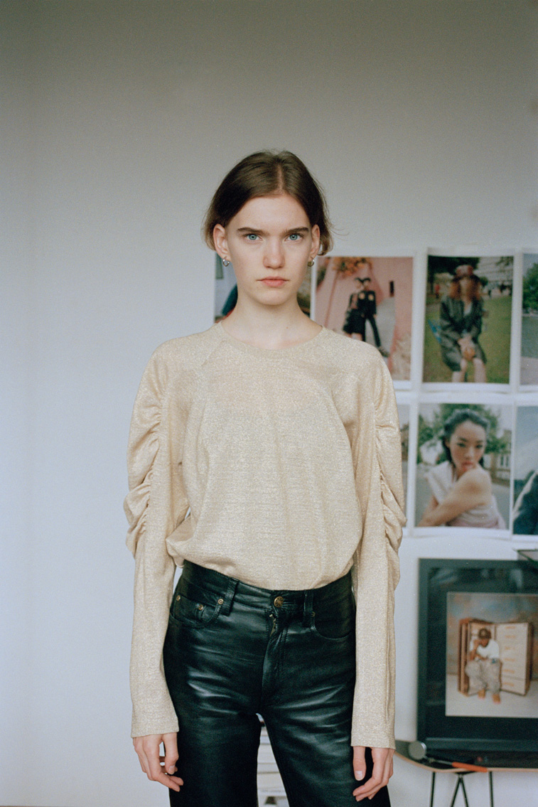 a young model in leather-look jeans and a gold lurex top stands looking to camera against a wall of photos