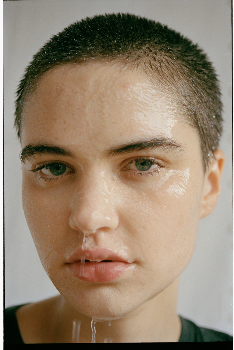 an androgynous model with a shaved head looks to camera with water dripping from their face