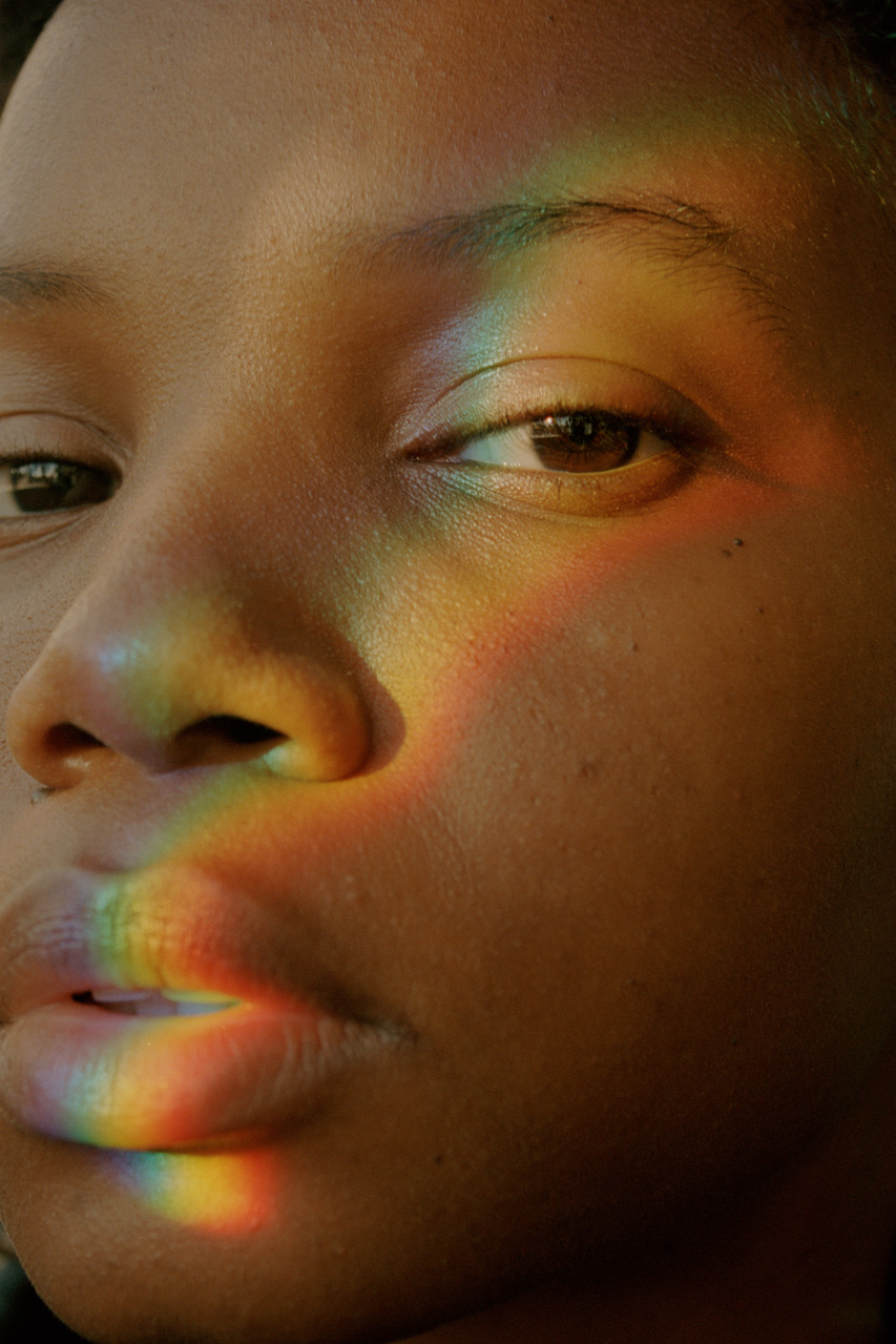 a close up of a Black model's face with a rainbow of light across their eye, cheek and lips