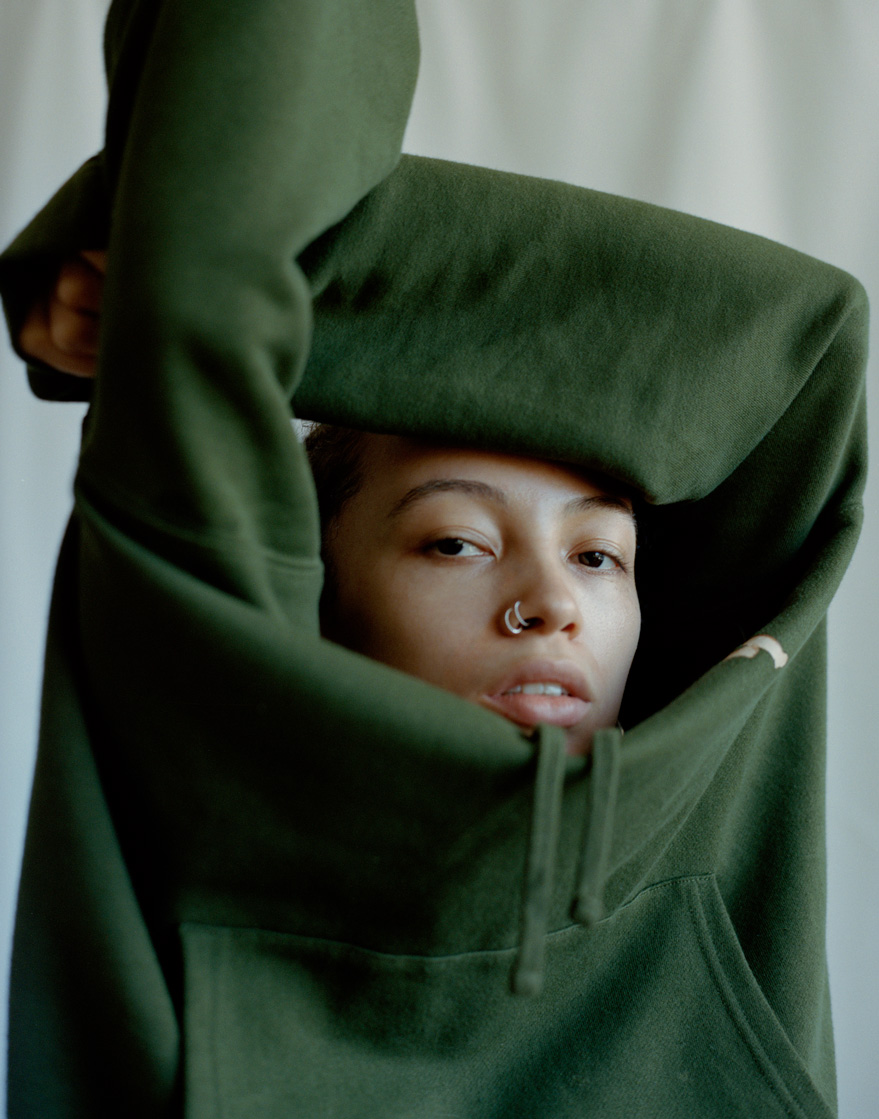 a mixed-race model with a nose piercing wearing a green hoodie holds her arms above her head