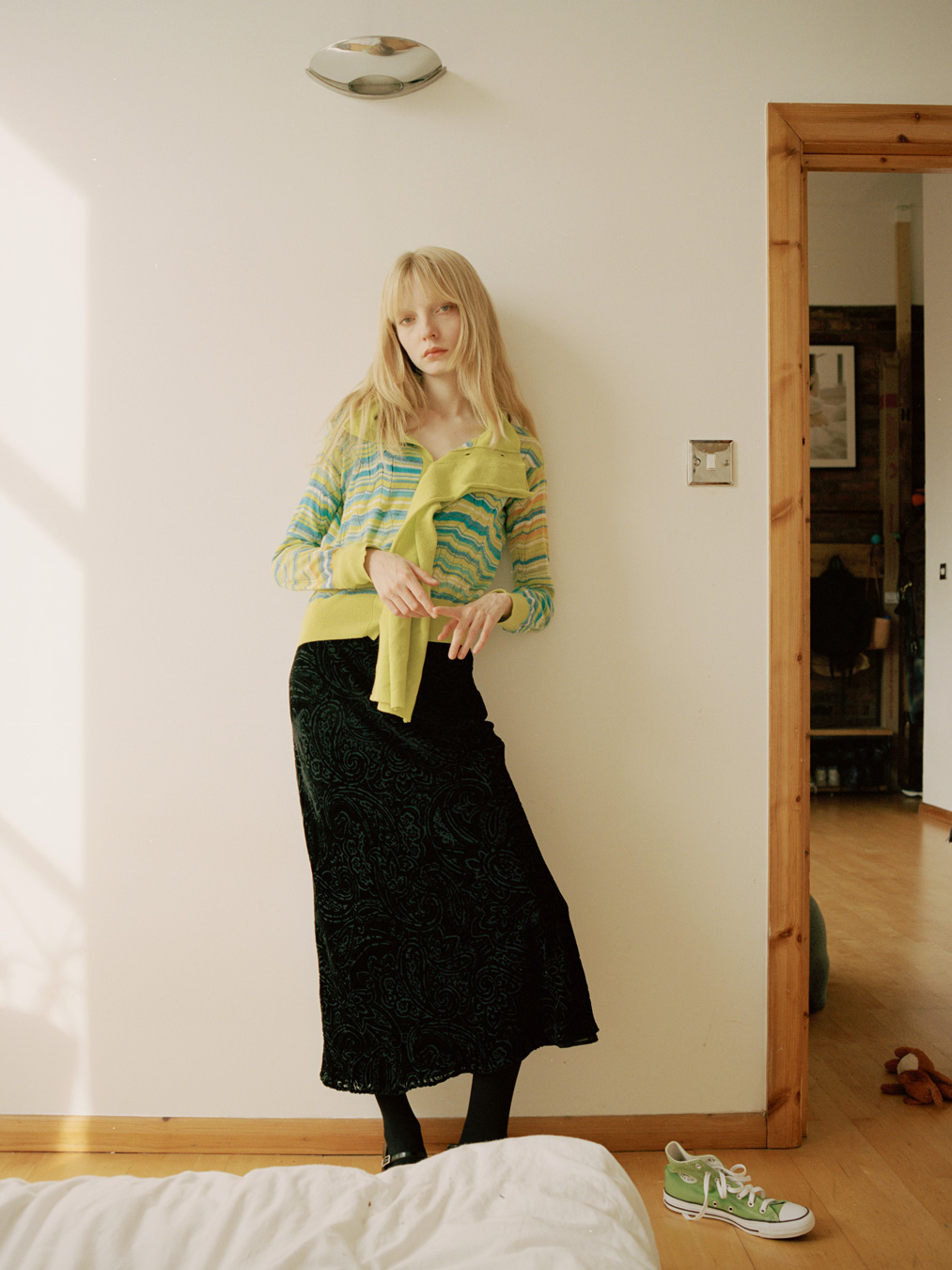 a blonde model in a long black dress and striped cardigan leans casually against a bedroom wall in the sun