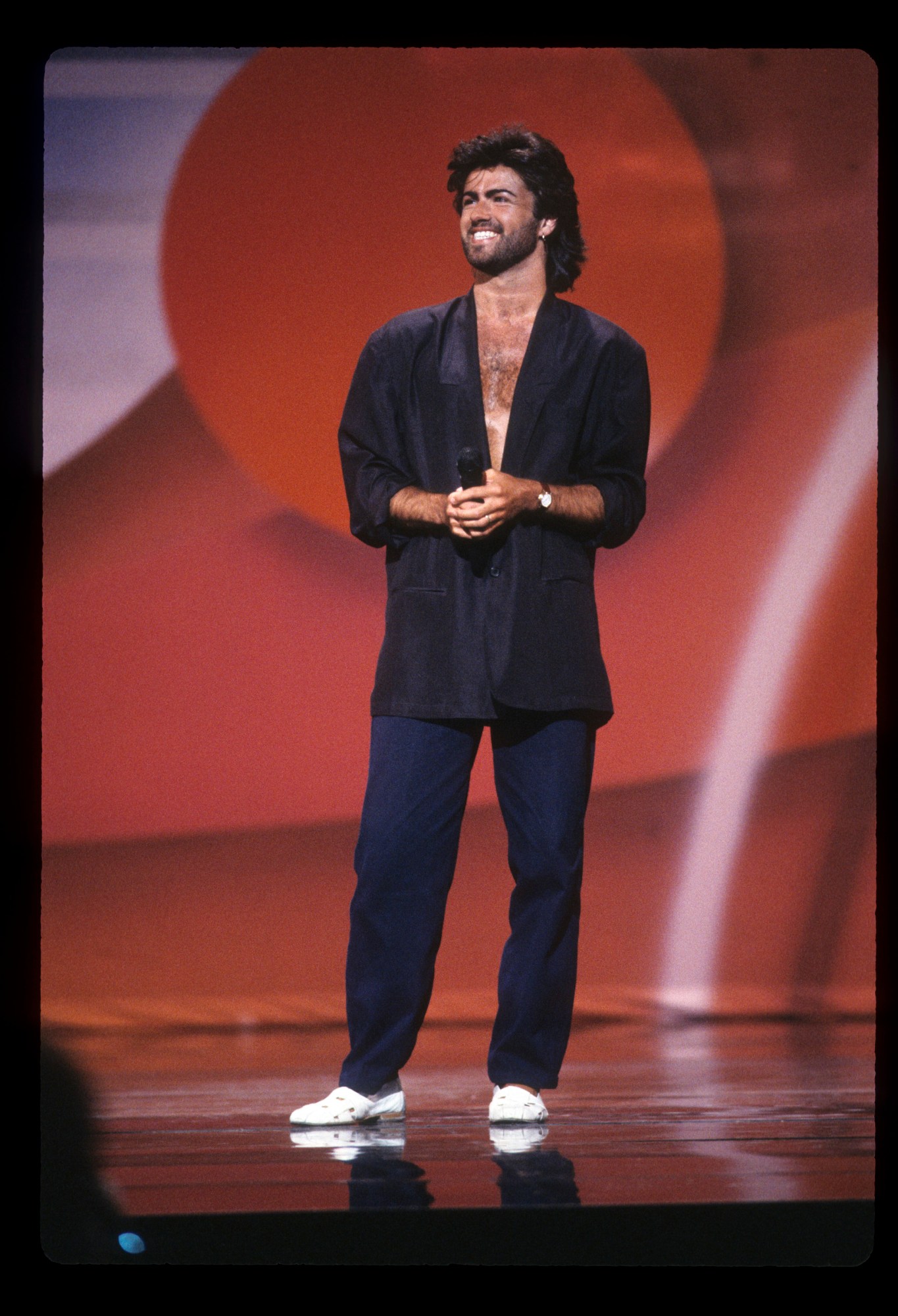 george michael on stage in dark trousers and an oversized blazer with a bare chest