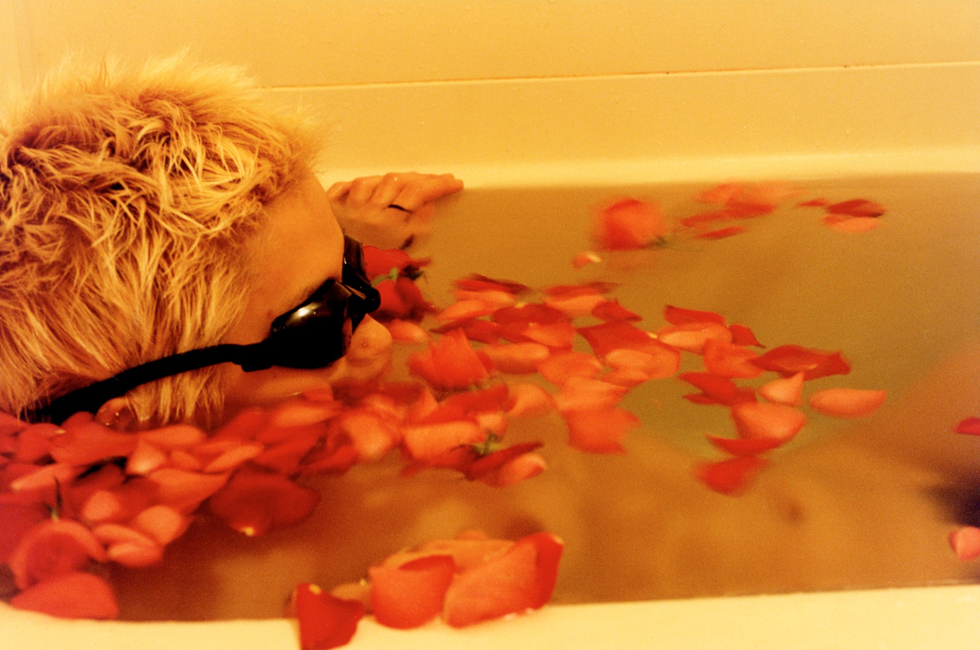 woman with short blonde hair lying in a bath wearing goggles covered with rose petals