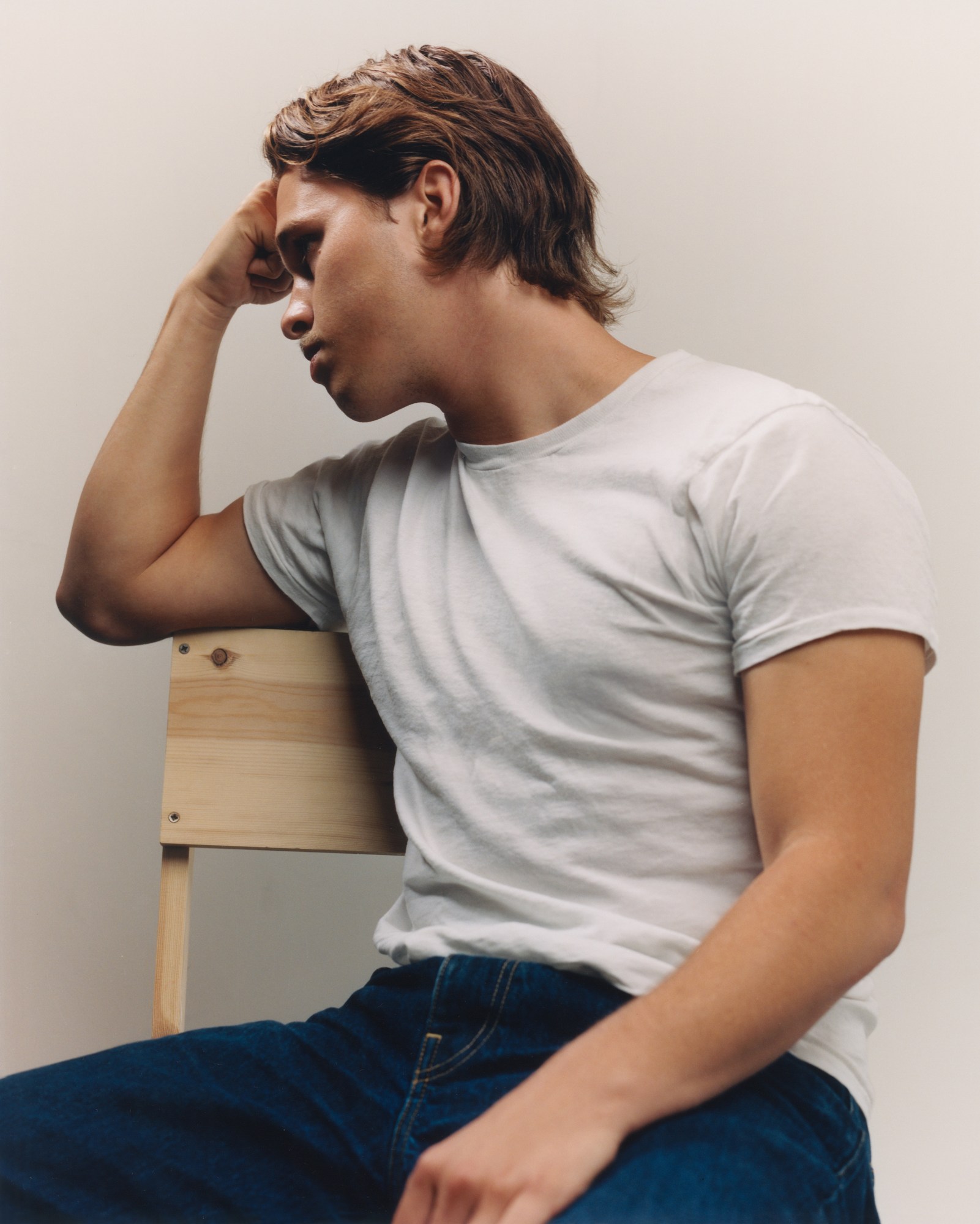 a portrait of the musician ryan beatty. he is a white man in his mid-twenties with a moustache and blonde hair. he's wearing a white t-shirt and blue-jeans. he's sat on a wooden chair resting his arm against it and his fist on his forehead, looking off to his right.