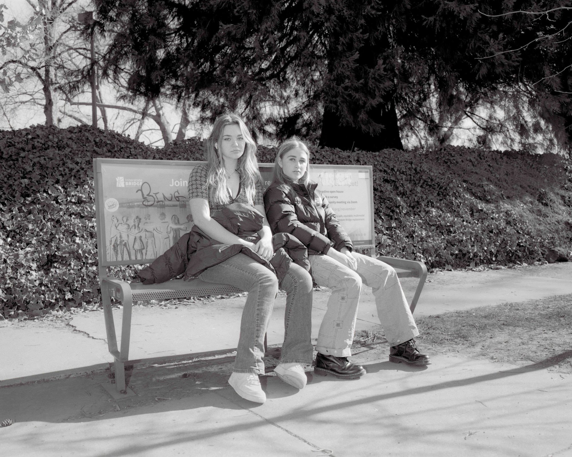 two young girls wearing jeans and puffer jackets sitting on a bench